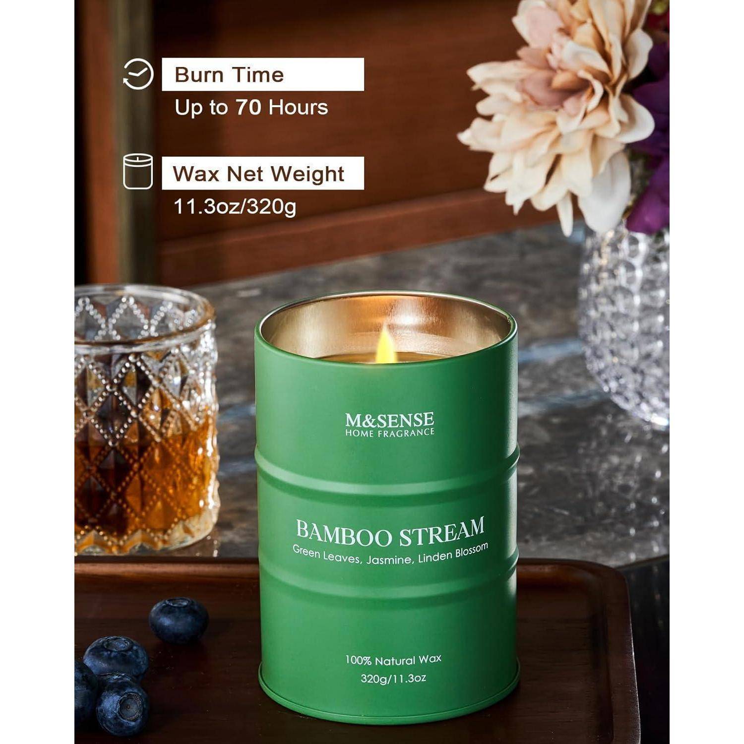 Eine grüne duftende Kerze mit der Aufschrift ‚Bamboo Stream' steht auf einem Holztablett mit Blumen und einem Glas in der Nähe. Text: ‚Brenndauer bis zu 70 Stunden, Wachsnetto-Gewicht 11,3 oz/320 g.