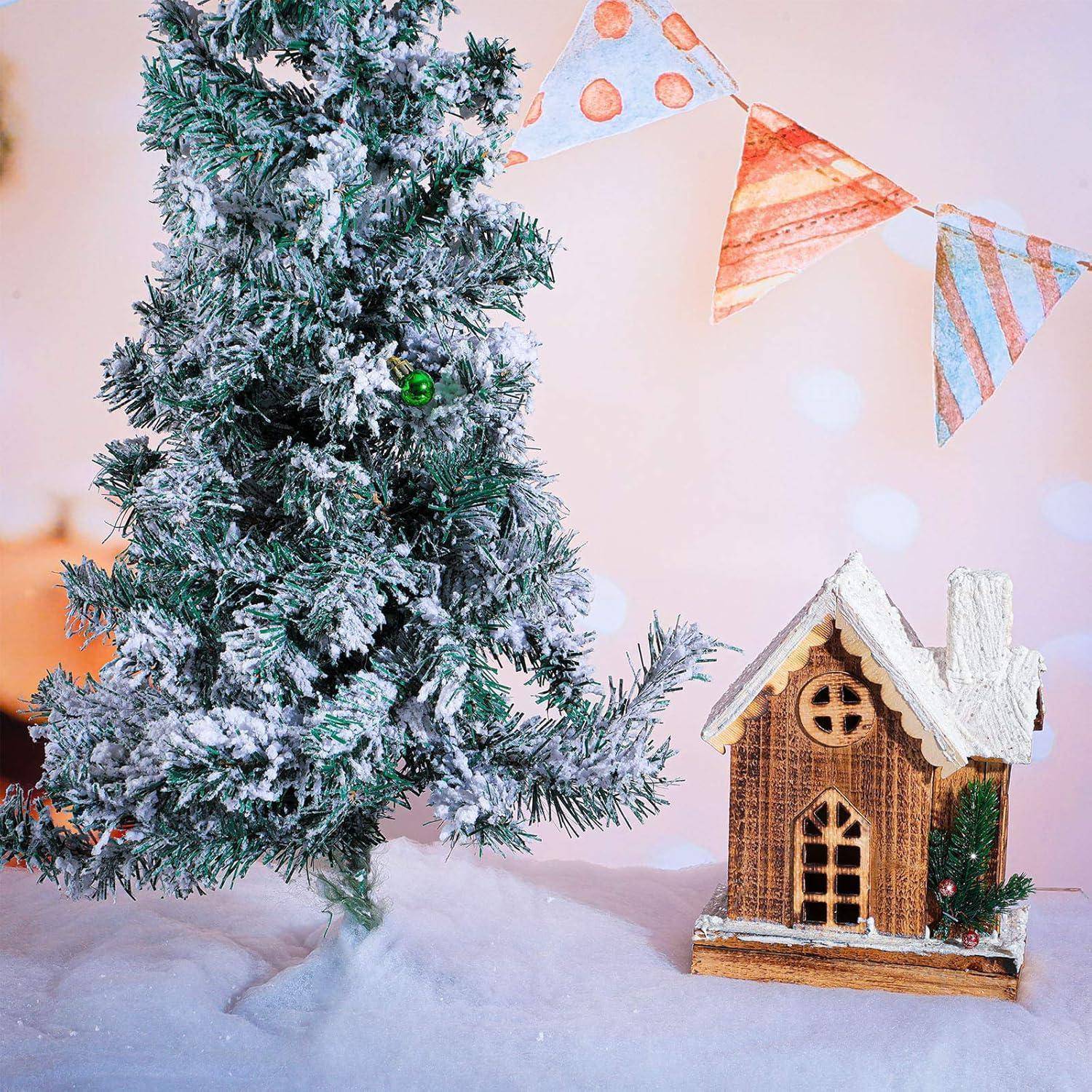 Eine kleine, schneebedeckte Kiefer steht neben einem Holzhausmodell, vor einem pastellfarbenen Hintergrund mit festlicher Wimpelkette.