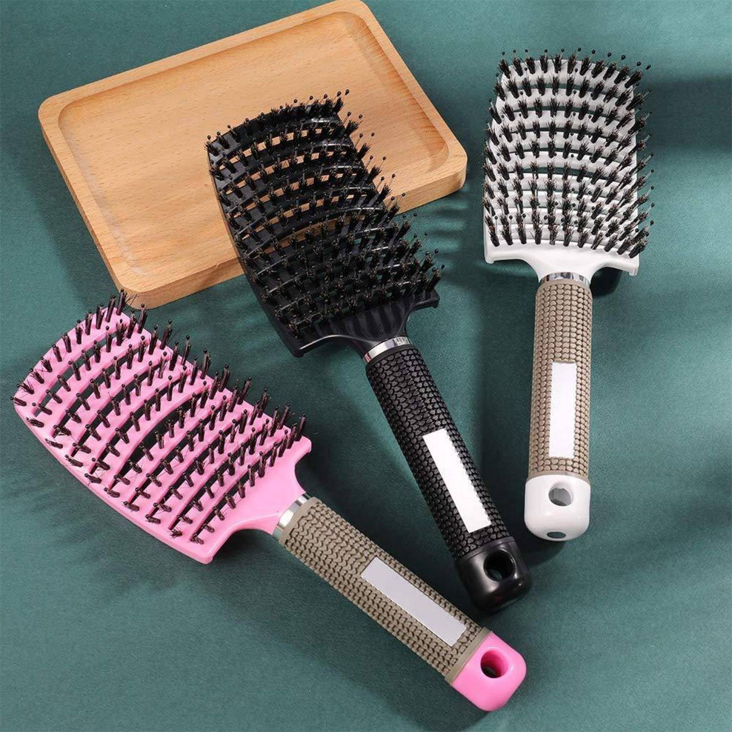 Drei belüftete Haarбürsten mit unterschiedlich farbigen Griffen (rosa, schwarz, braun) liegen auf einer grünen Oberfläche neben einem Holztablett.