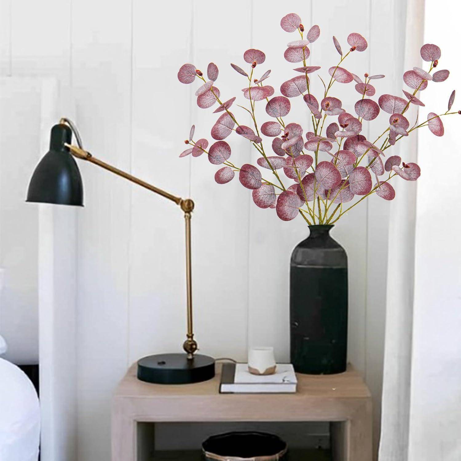 Ein Schreibtisch mit einer schwarzen Lampe, Büchern und einer großen Vase mit rosa Eukalyptusblättern in einem modernen Raumambiente.
