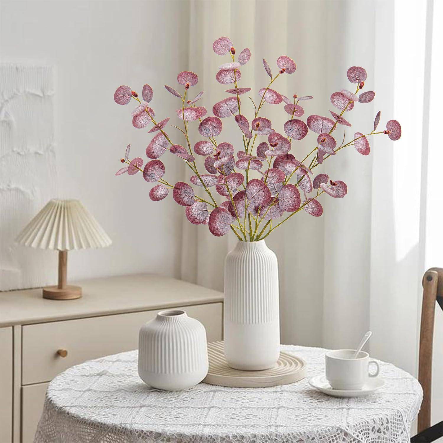 Ein Blumenvase mit pinken Blumen auf einem Tisch, begleitet von einem kleinen Topf und einer Tasse. Eine Lampe steht auf einem Holzschrank im Hintergrund.