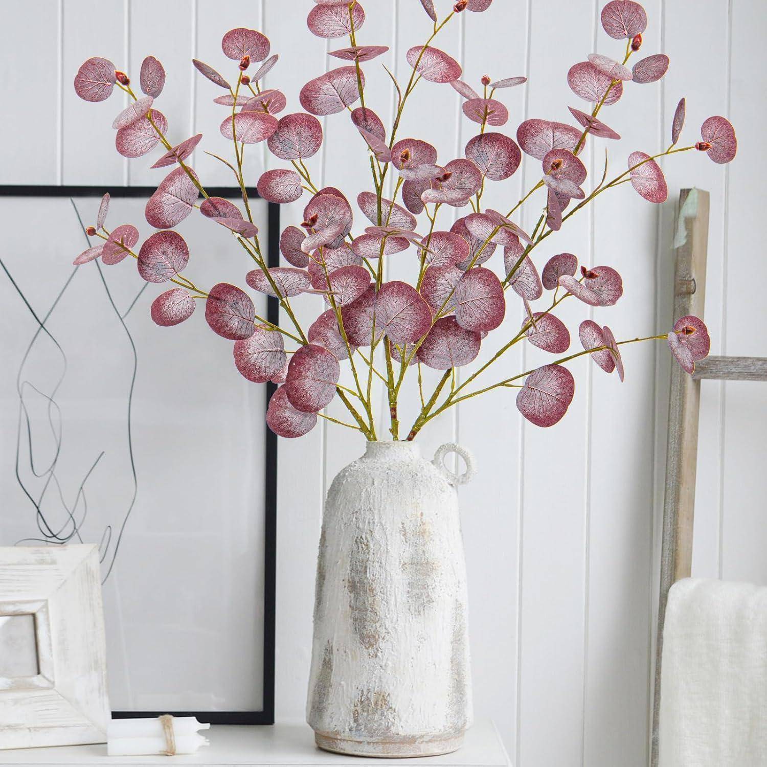 Eine weiße Vase mit rosa Eukalyptuszweigen steht auf einem weißen Regal, neben einem minimalistischen Linienkunstgemälde und Büchern.