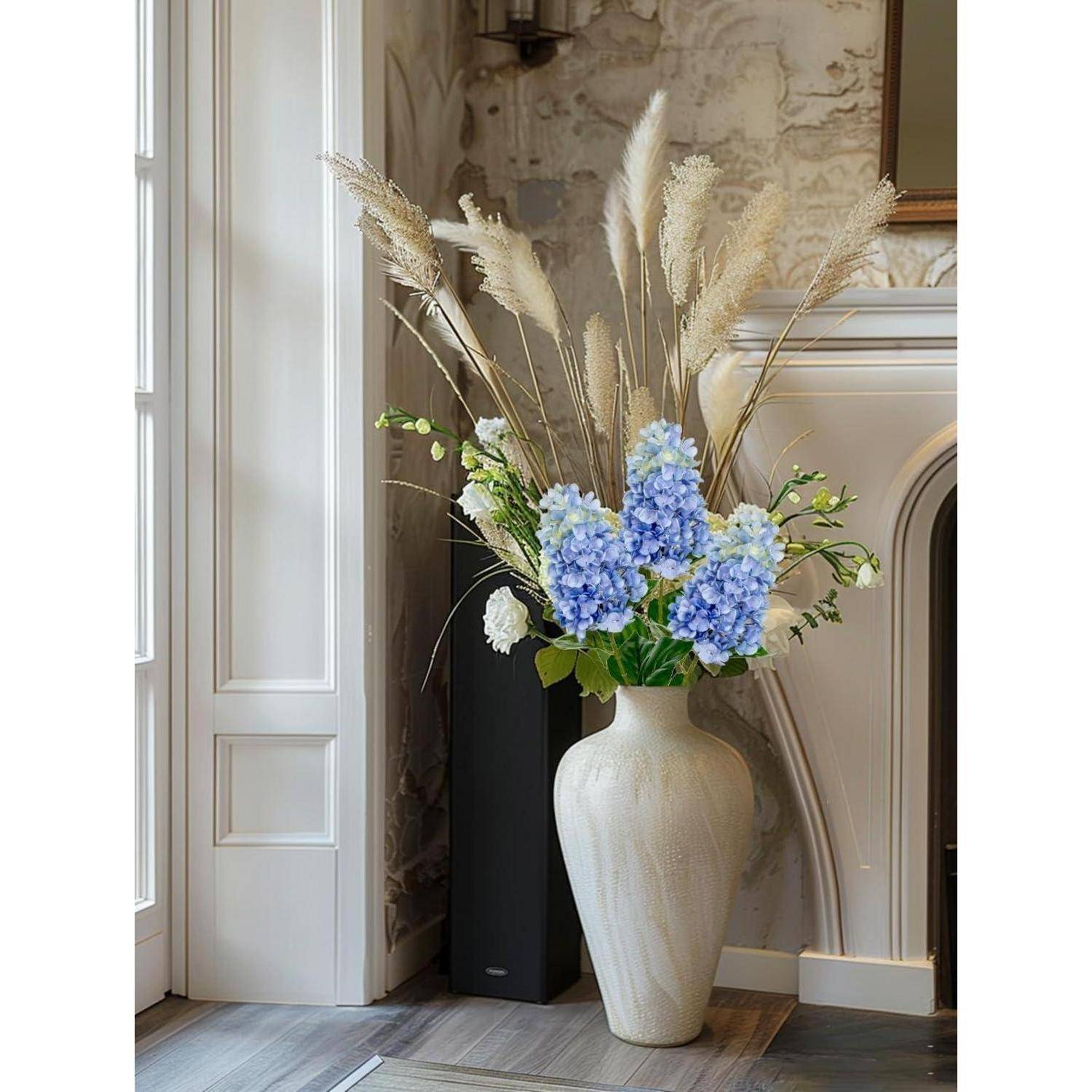 Eine beige Keramikvas mit getrockneten Gräsern und blauen Hortensien steht neben einem Kamin in einem gemütlichen Raum mit elegantem Dekor.