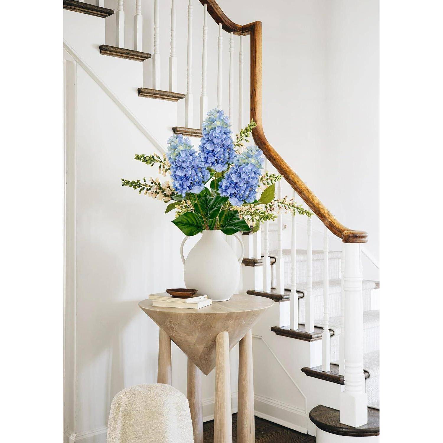 Weiße Treppe mit einem Holzgeländer, blaue Hortensien in einer weißen Vase auf einem kleinen Tisch am Fuß der Treppe und einem plüschigen Hocker in der Nähe.