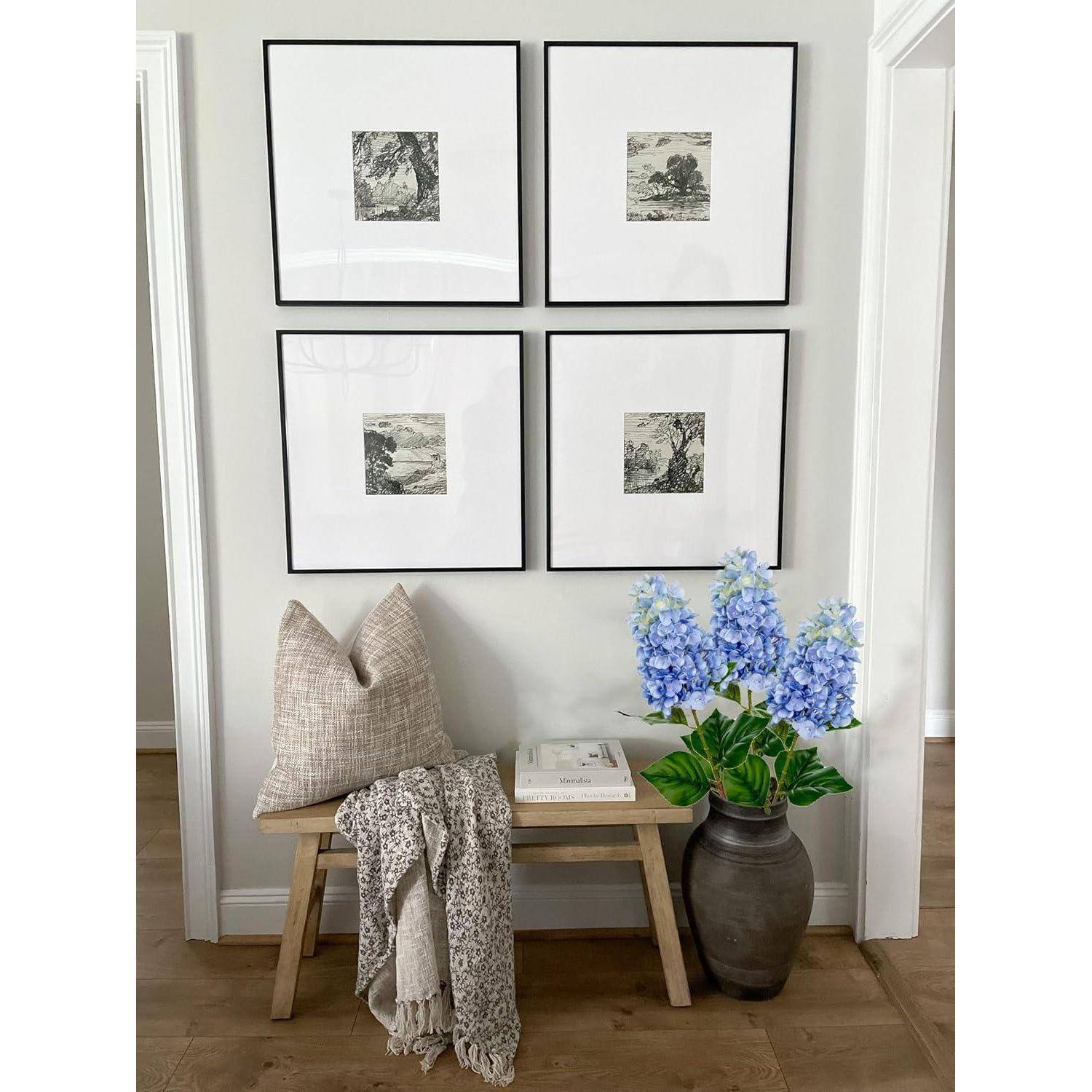 Ein minimalistischer Flur zeigt eine Holzbank mit einem Kissen und einer drappierten Decke. Eine Vase mit blauen Blumen steht neben vier gerahmten Kunstwerken.