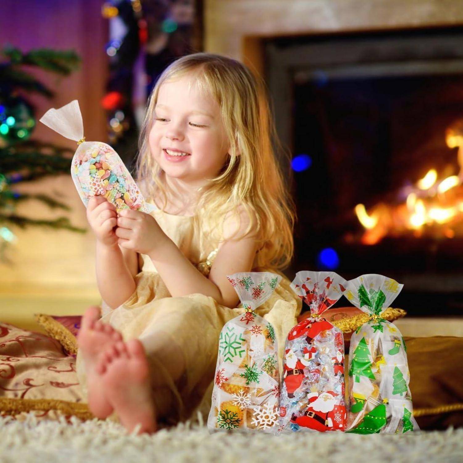 Ein kleines Kind lächelt, während es eine Geschenktüte hält und nahe einem Kamin sitzt, der mit festlichen Dekorationen geschmückt ist und von weiteren Geschenktüten umgeben ist.
