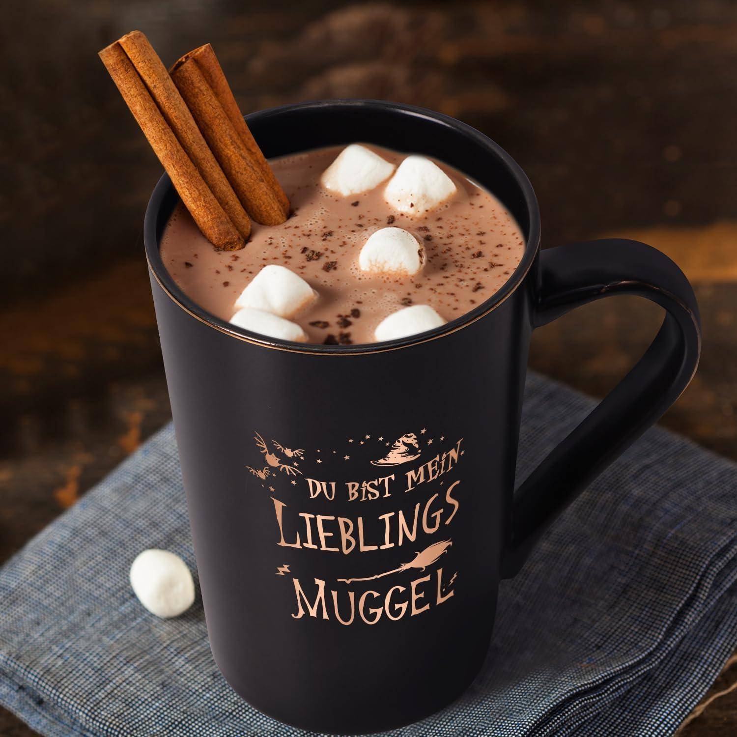Eine schwarze Tasse gefüllt mit heißer Schokolade, gekrönt von Marshmallows und Zimtstangen, steht auf einem blauen Tuch. Die Tasse zeigt deutschen Text.