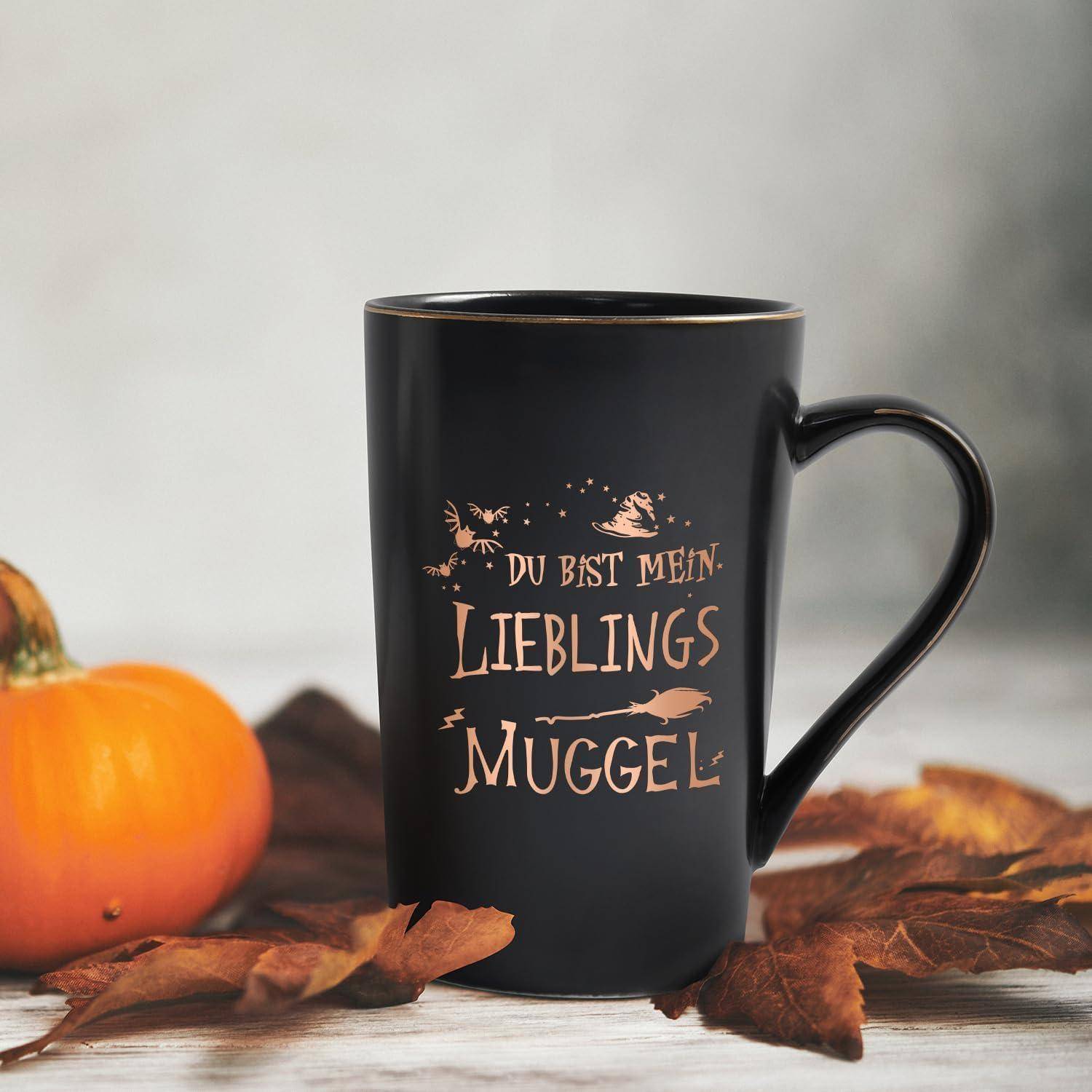 Eine schwarze Tasse mit goldener Aufschrift ‚Du bist mein Lieblings Muggel' liegt zwischen Herbstblättern und einem kleinen Kürbis und schafft eine gemütliche Herbststimmung.