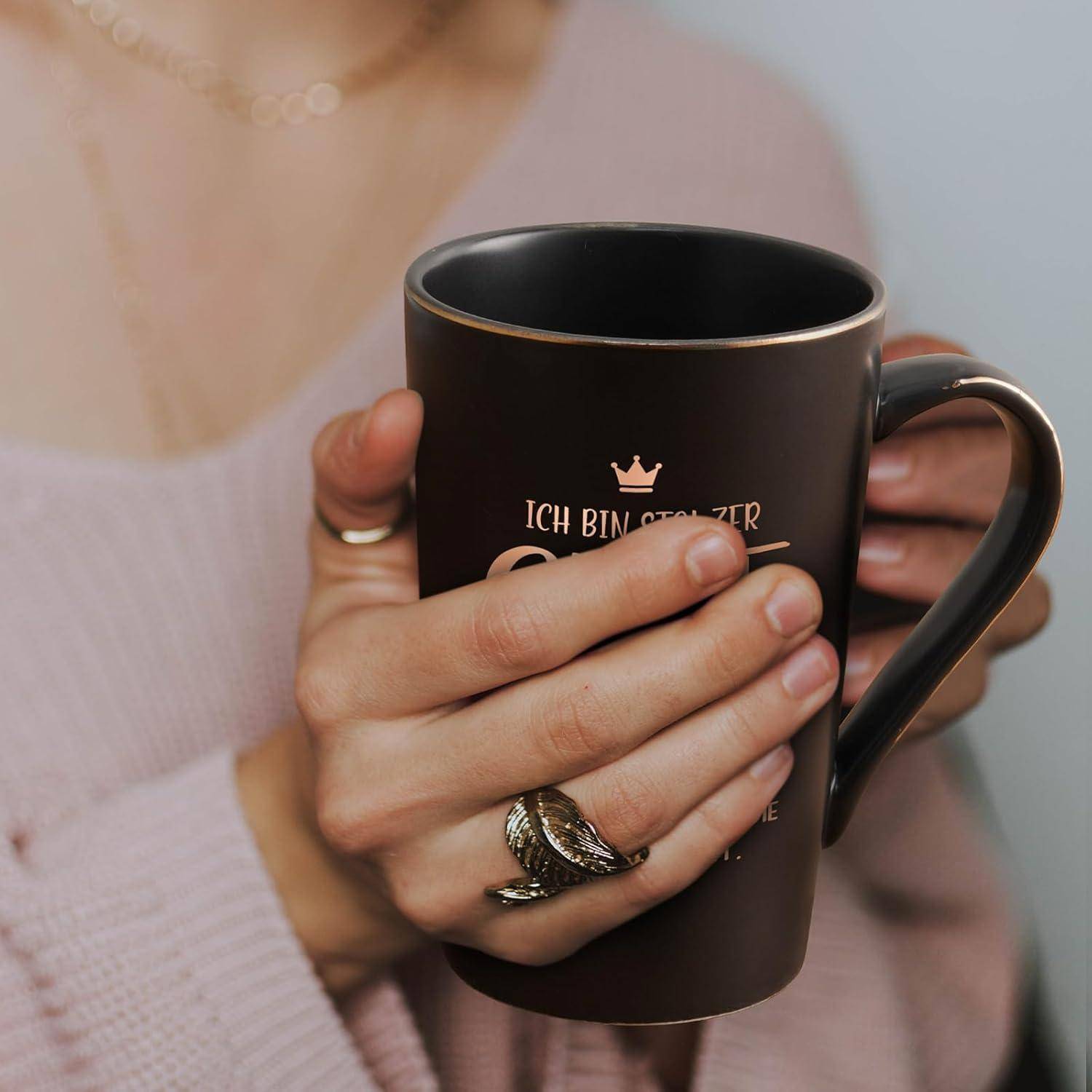 Eine Person, die eine schwarze Tasse mit goldener Schrift hält. Der Text lautet 