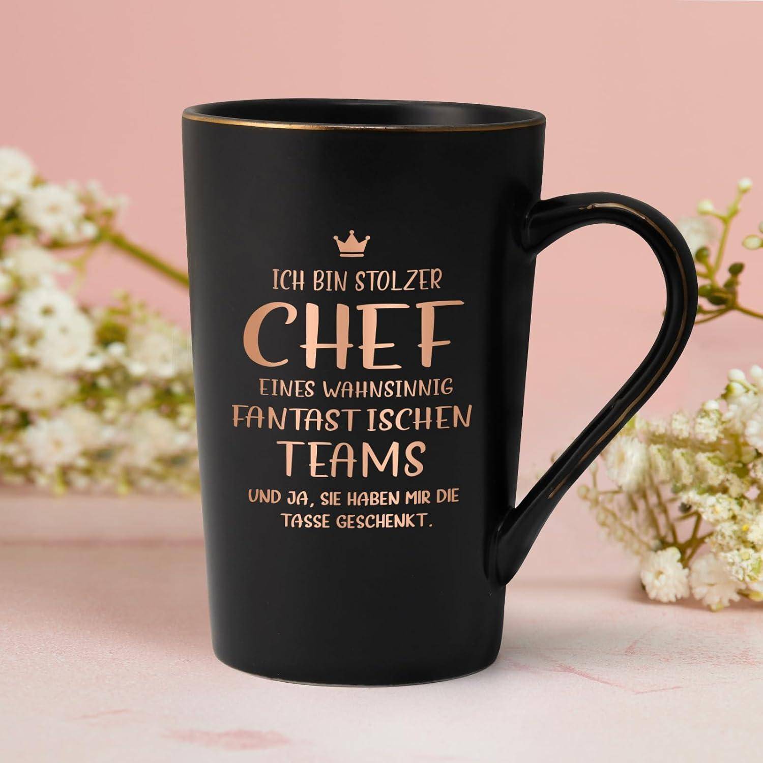 Ein schwarzer Becher mit goldener Schrift in Deutsch liest sich: 'Ich bin stolzer Chef eines wahnsinnig fantastischen Teams und ja, sie haben mir die Tasse geschenkt.