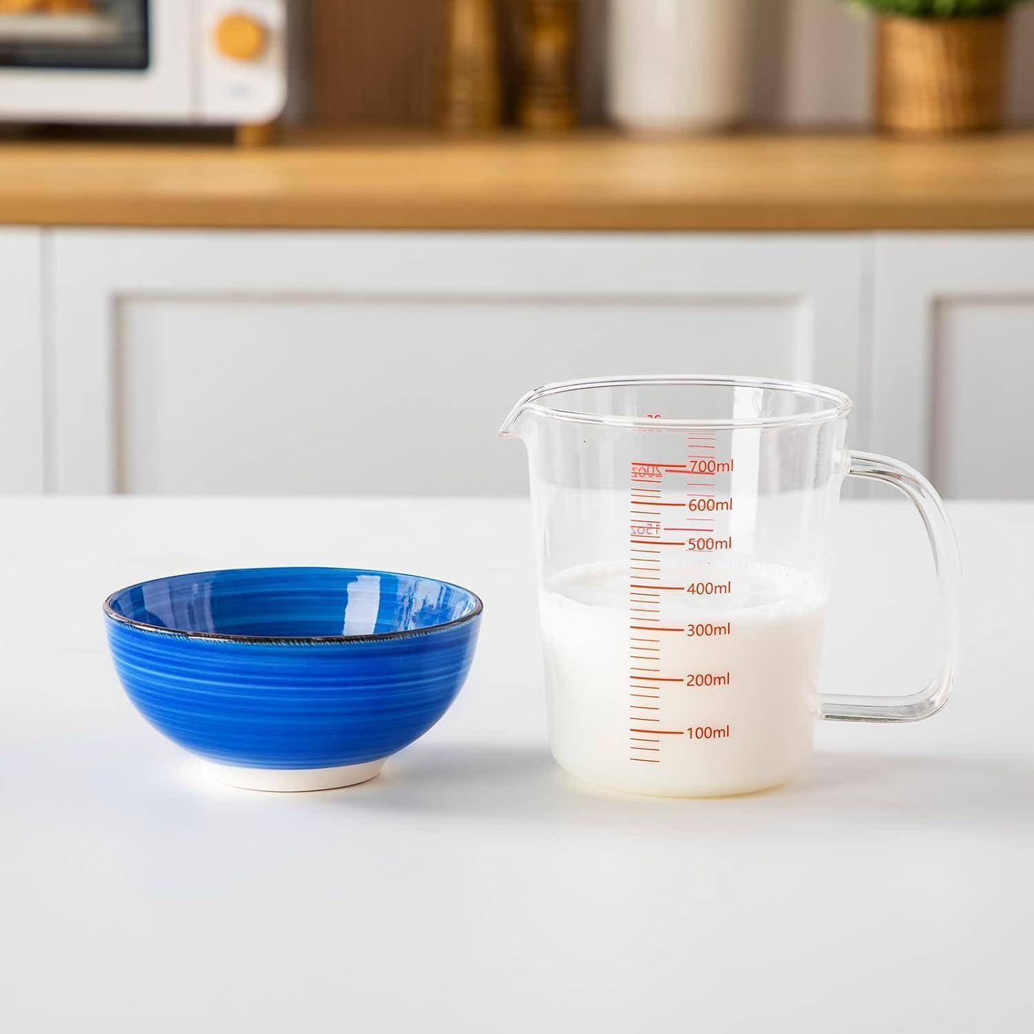 Eine durchsichtige Messbecher mit Milch steht neben einer blauen Schüssel auf einer weißen Küchenarbeitsplatte.
