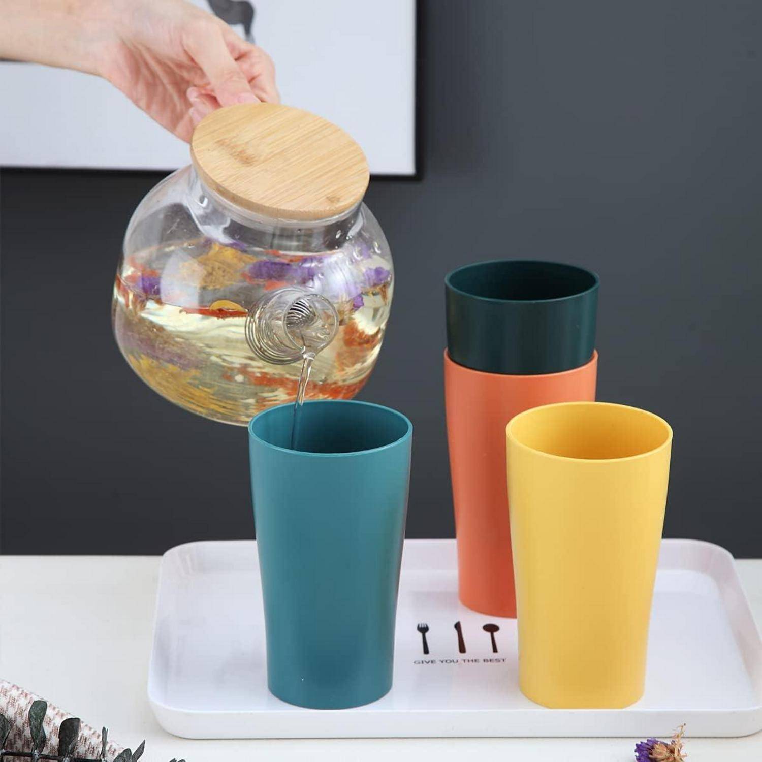 Eine Hand gießt Tee aus einer Glasteekanne in eine petrolblaue Tasse. Zwei weitere Tassen, eine in Orange und eine in Gelb, stehen auf einem weißen Tablett neben kleinen Löffeln.