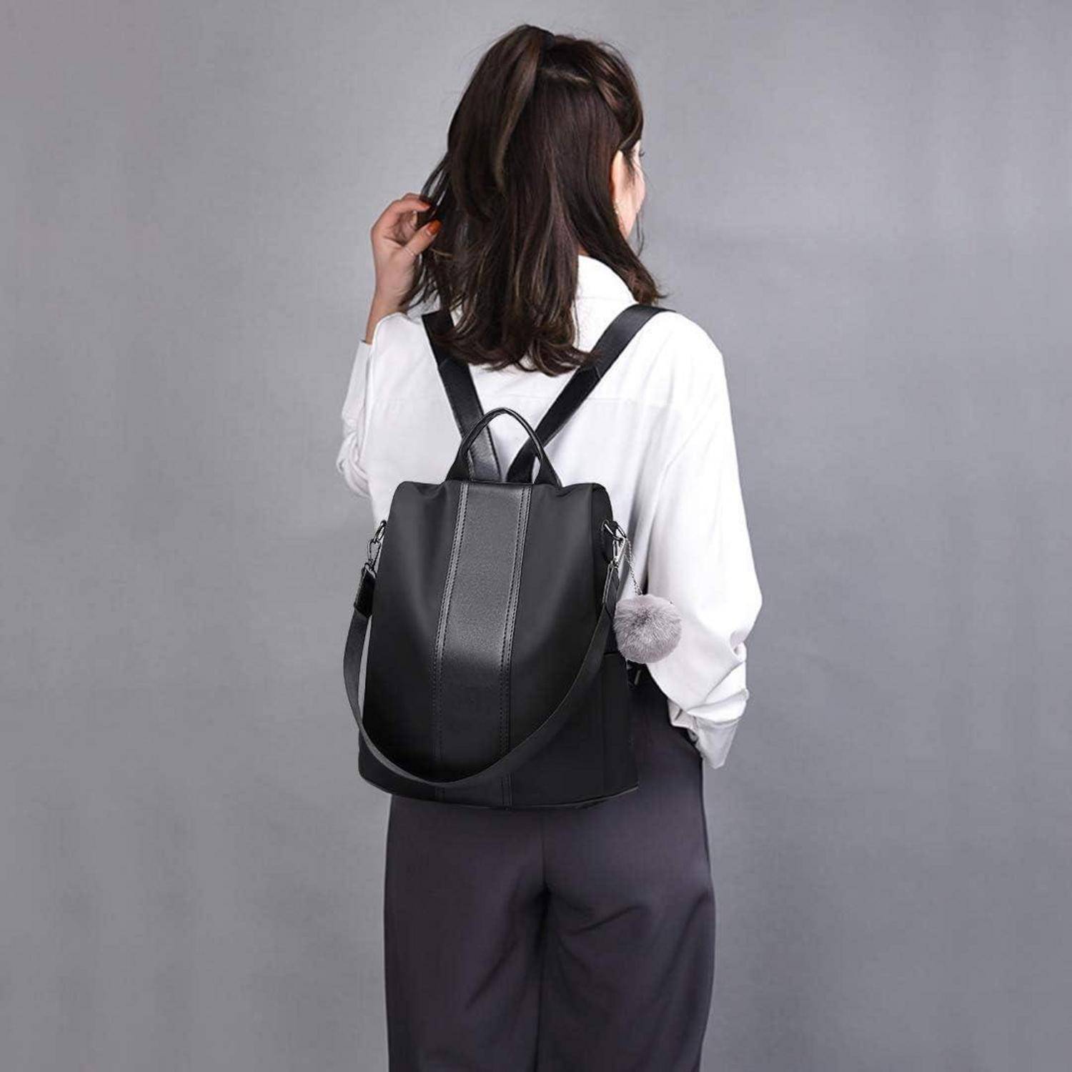 Eine Person mit langen Haaren, trägt ein weißes Hemd und schwarze Hose, von hinten zu sehen, während sie einen schwarzen Lederrucksack vor einem grauen Hintergrund trägt.