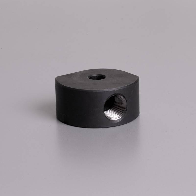 Ein kleines, schwarzes, zylindrisches Metallstück mit einem Gewindeloch an seiner Seite, platziert auf einer einfachen, hellgrauen Oberfläche.