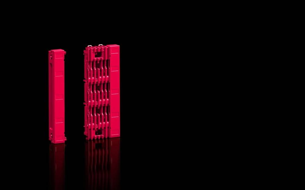 Zwei hohe, rote vertikale Strukturen mit komplexen, sich wiederholenden Mustern stehen auf einer glänzenden schwarzen Oberfläche, deren Formen sich darin spiegeln.