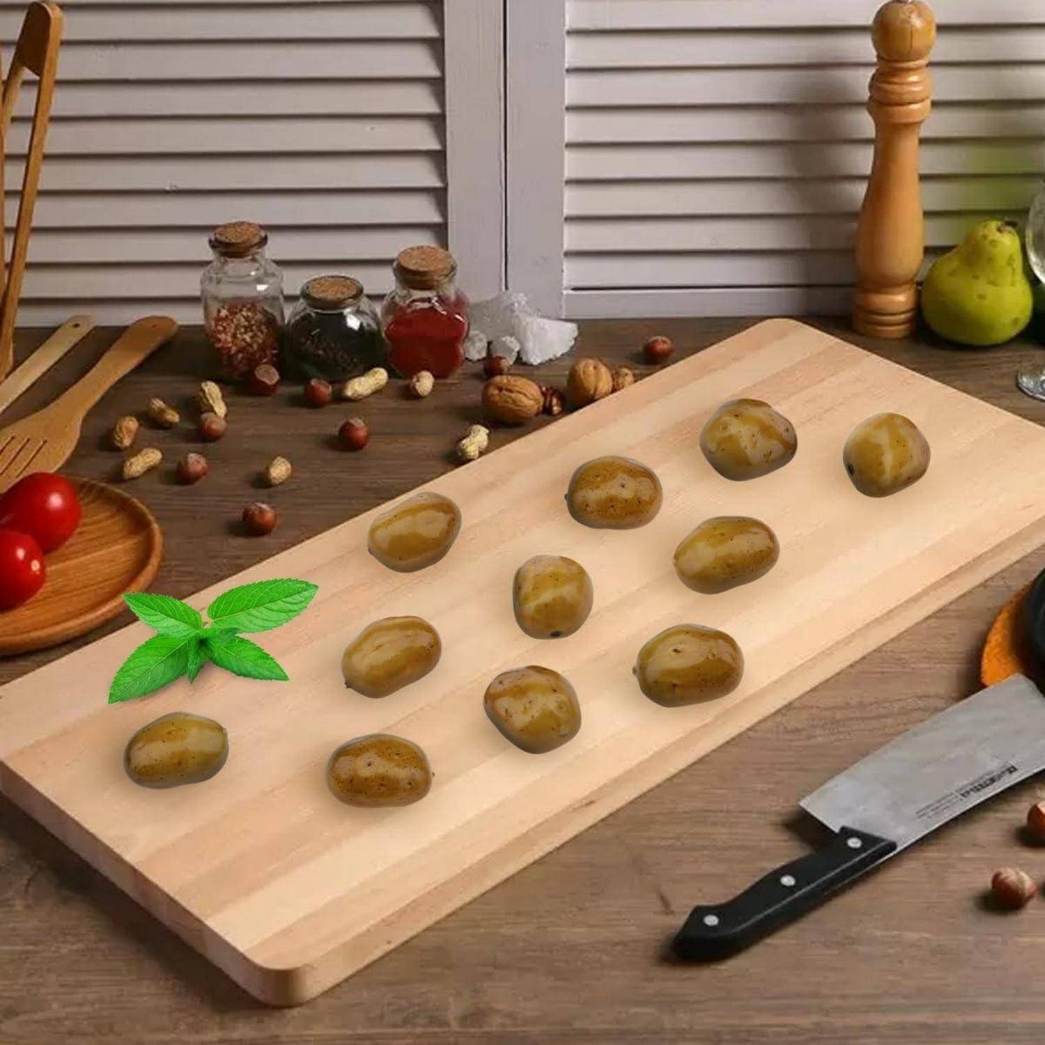 Ein Holzschneidebrett mit kleinen Kartoffeln, einem Messer und Basilikumblättern auf einer Küchenarbeitsplatte. Verschiedene Gewürze und Früchte im Hintergrund.