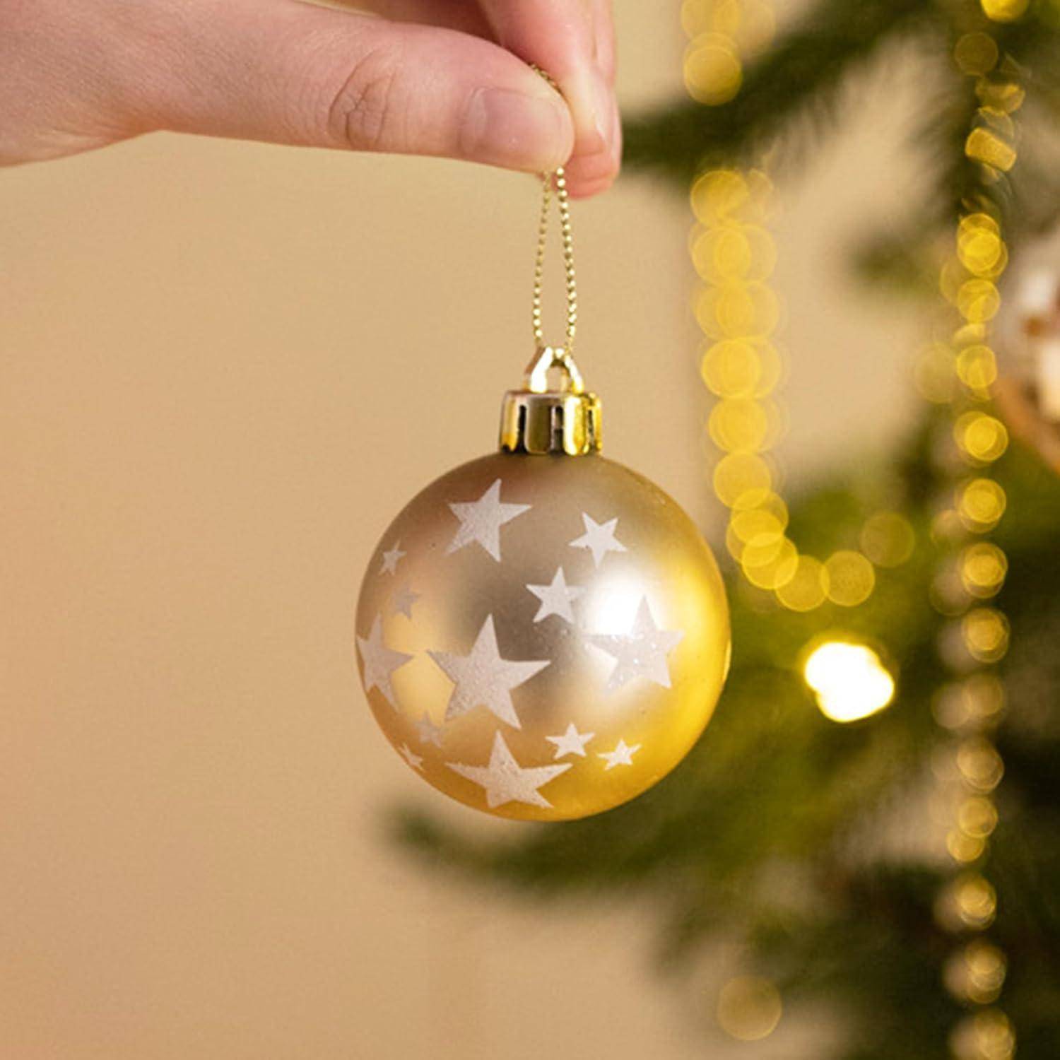 Eine Hand hält eine goldene Weihnachtskugel mit weißen Sternenmustern, vor einem unscharfen Weihnachtsbaum mit leuchtend gelben Lichtern.