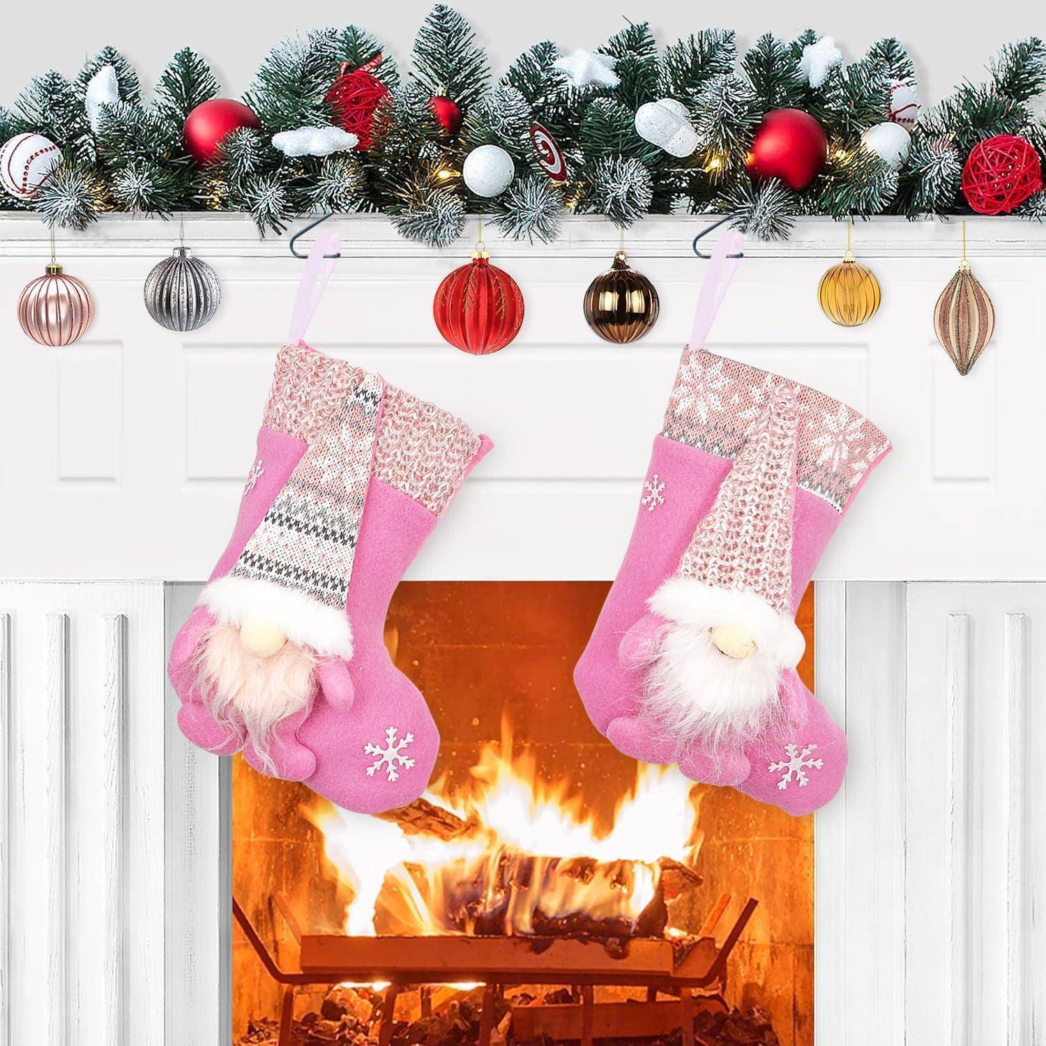 Festlicher Kamin mit Weihnachtsstrümpfen: Zwei rosa Weihnachtsstrümpfe hängen über einem mit Girlande und Weihnachtskugeln geschmückten Kaminsims. Darunter lodert ein helles Feuer.