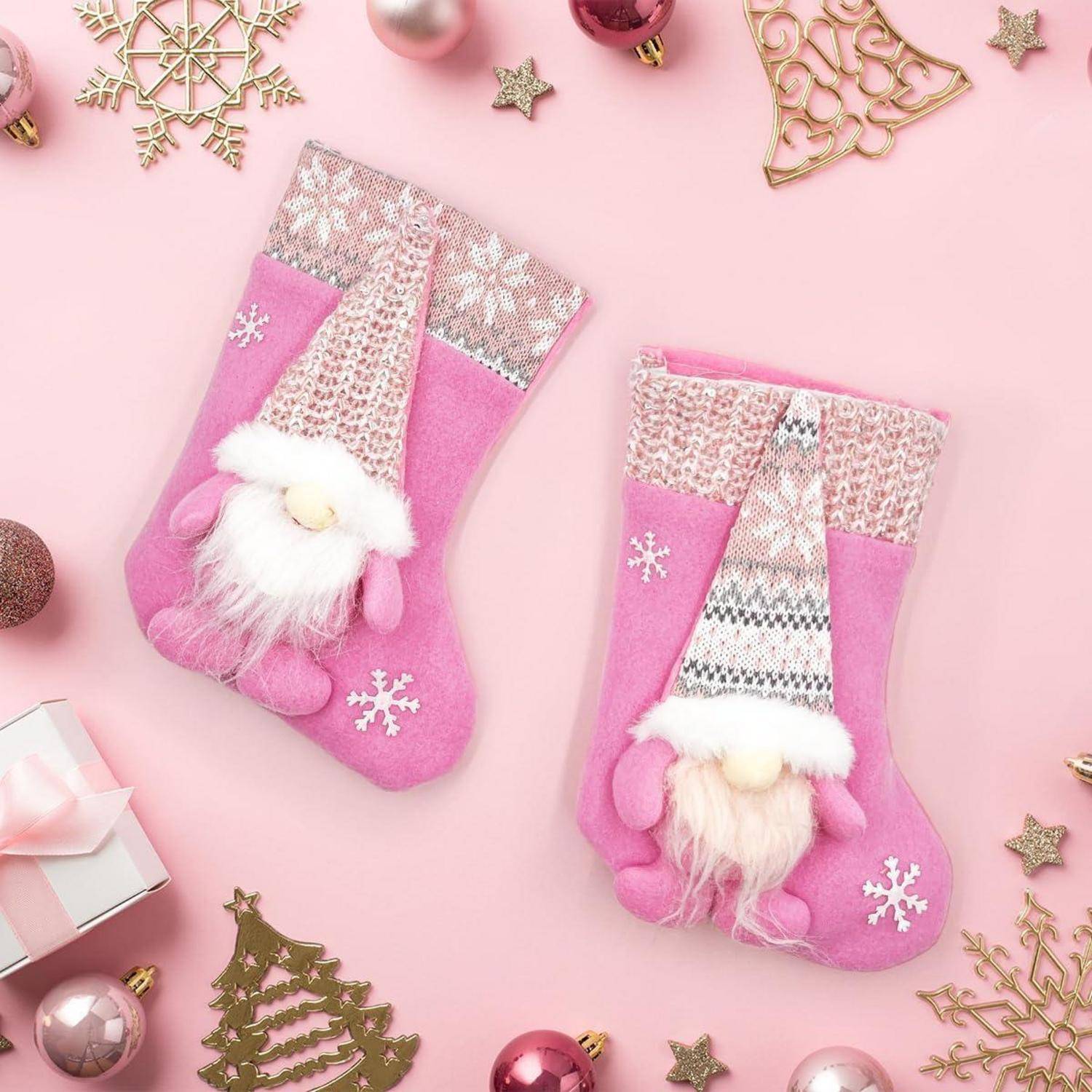Zwei rosa Weihnachtssocken mit Wichteldesigns liegen auf einem rosa Hintergrund, umgeben von Ornamenten wie Schneeflocken und einer Geschenkbox.