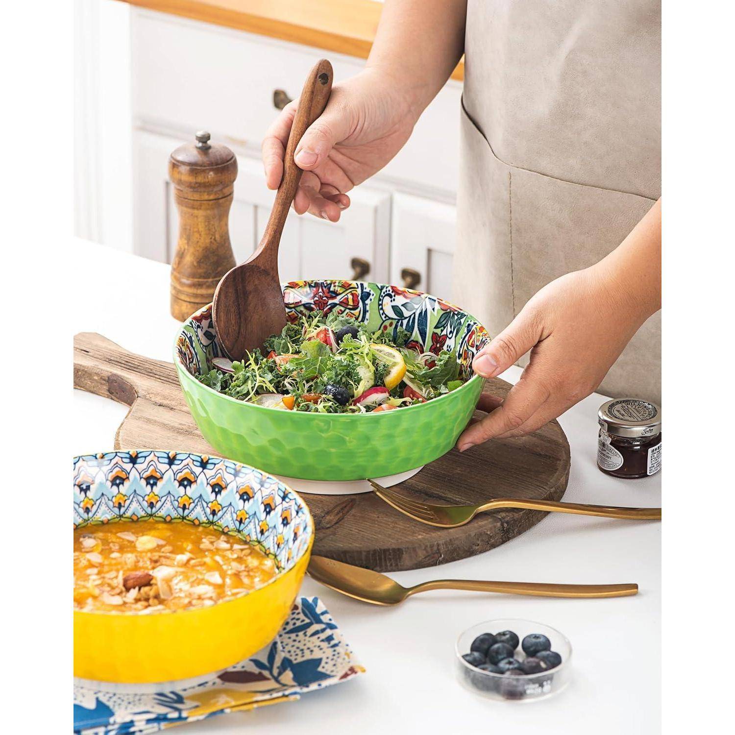 Eine Person serviert Salat aus einer bunten Schüssel mit Holzutensilien. Eine Suppenschüssel und Blaubeeren befinden sich daneben auf der Küchenarbeitsfläche.