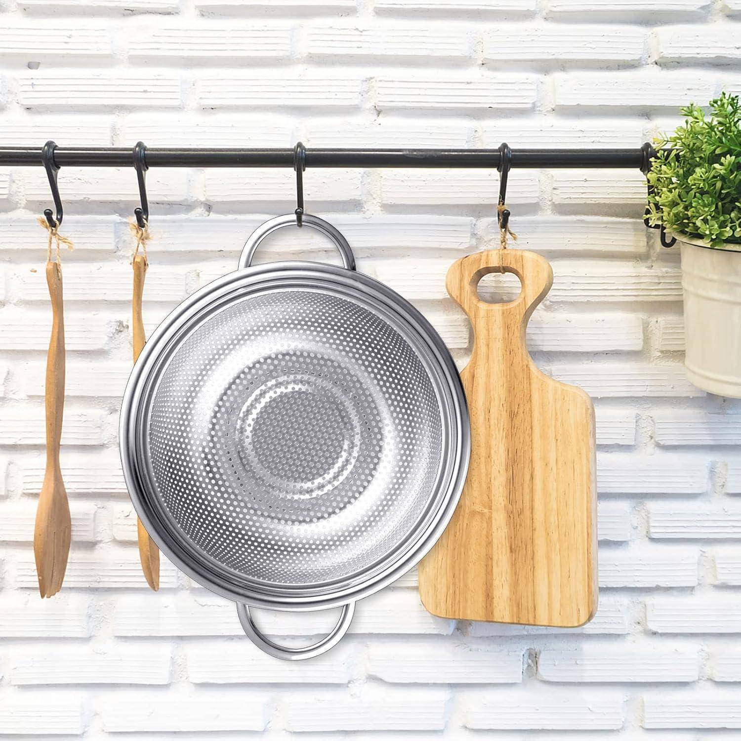 Eine Küchenwand mit einem hängenden Metalldurchschlag, einem Holzlöffel, einem Schneidebrett und einer kleinen Topfpflanze vor einem weißen Ziegelwandhintergrund.