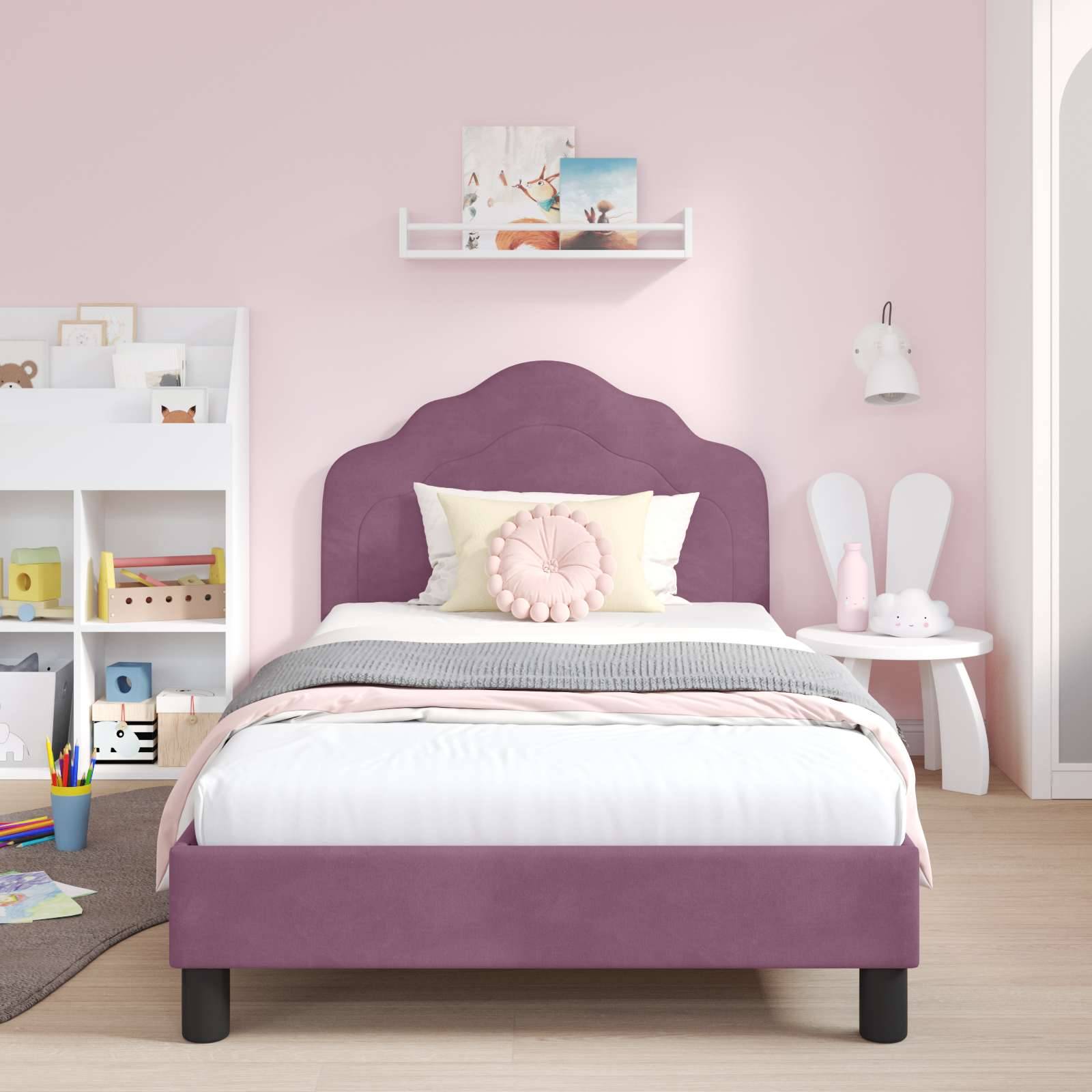 Ein rosa getöntes Schlafzimmer mit einem lila Bett, grauer Decke und rosa Kissen. Ein weißes Regal trägt Spielzeug und Bücher. Ein kleiner Tisch und Stühle befinden sich rechts.
