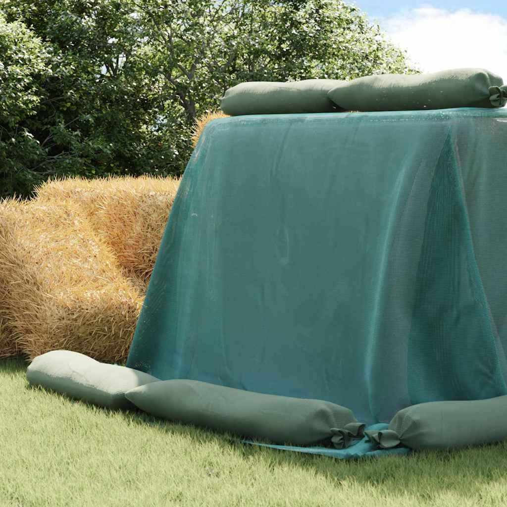 Ein Stapel Strohballen, der mit einer grünen Plane abgedeckt und von Sandsäcken umgeben ist, steht auf einem Grasfeld mit Bäumen im Hintergrund.