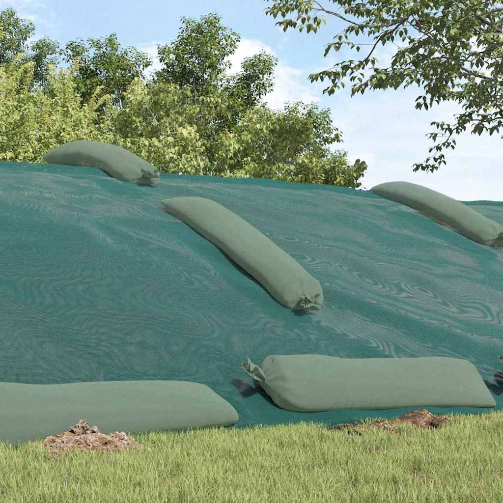 Ein mit Gras bewachsener Hang, der mit einer großen grünen Plane bedeckt ist, die durch Sandsäcke gesichert wird, unter einem klaren blauen Himmel und von Bäumen umgeben.