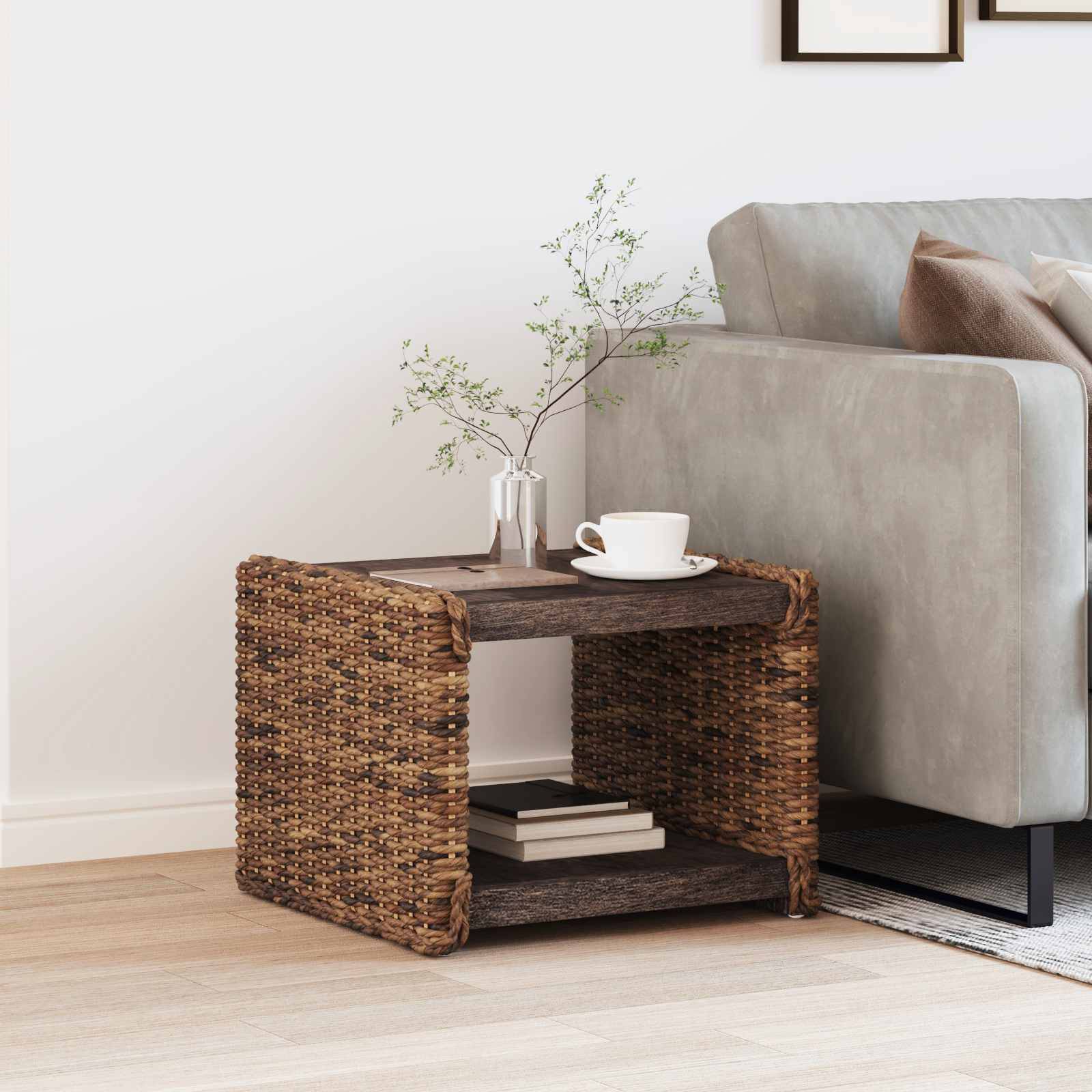 Ein modernes Wohnzimmer mit einem grauen Sofa, beigen Kissen und einem geflochtenen Beistelltisch, auf dem eine kleine Pflanze und eine Tasse stehen, vor einer hellen Wand.