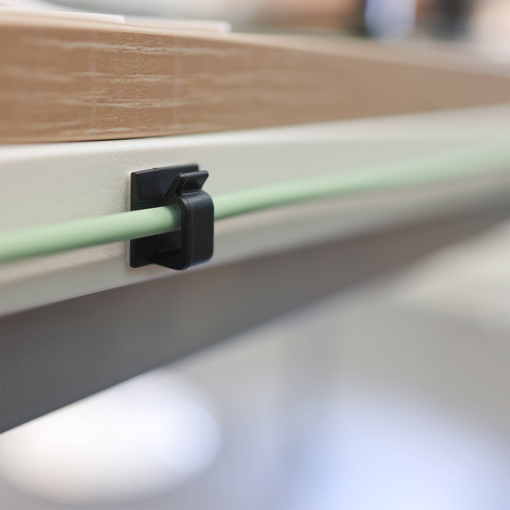 Detailaufnahme einer Kabelführungsklemme, die ein dünnes grünes Kabel an der Kante einer hellholzfarbenen Oberfläche befestigt.