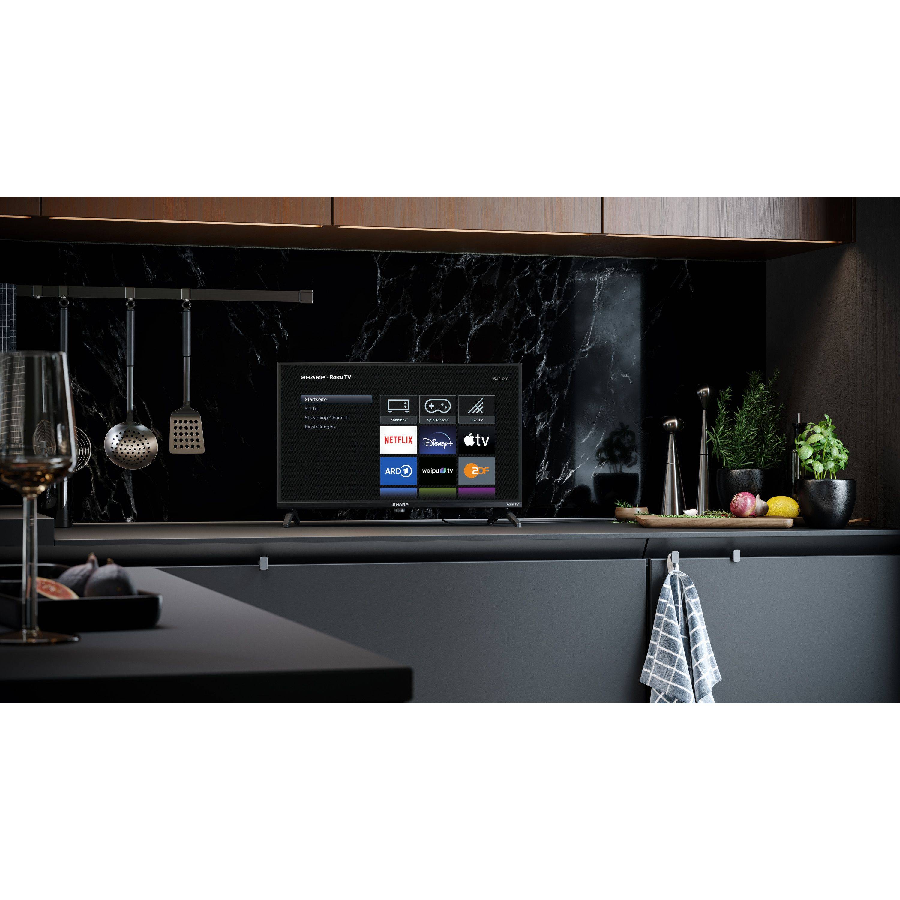 Eine elegante Küchenszene mit einem modernen schwarzen Fernseher auf der Arbeitsplatte, auf dem Streaming-App-Symbole wie Netflix und YouTube zu sehen sind. Ein Schneidebrett liegt in der Nähe.