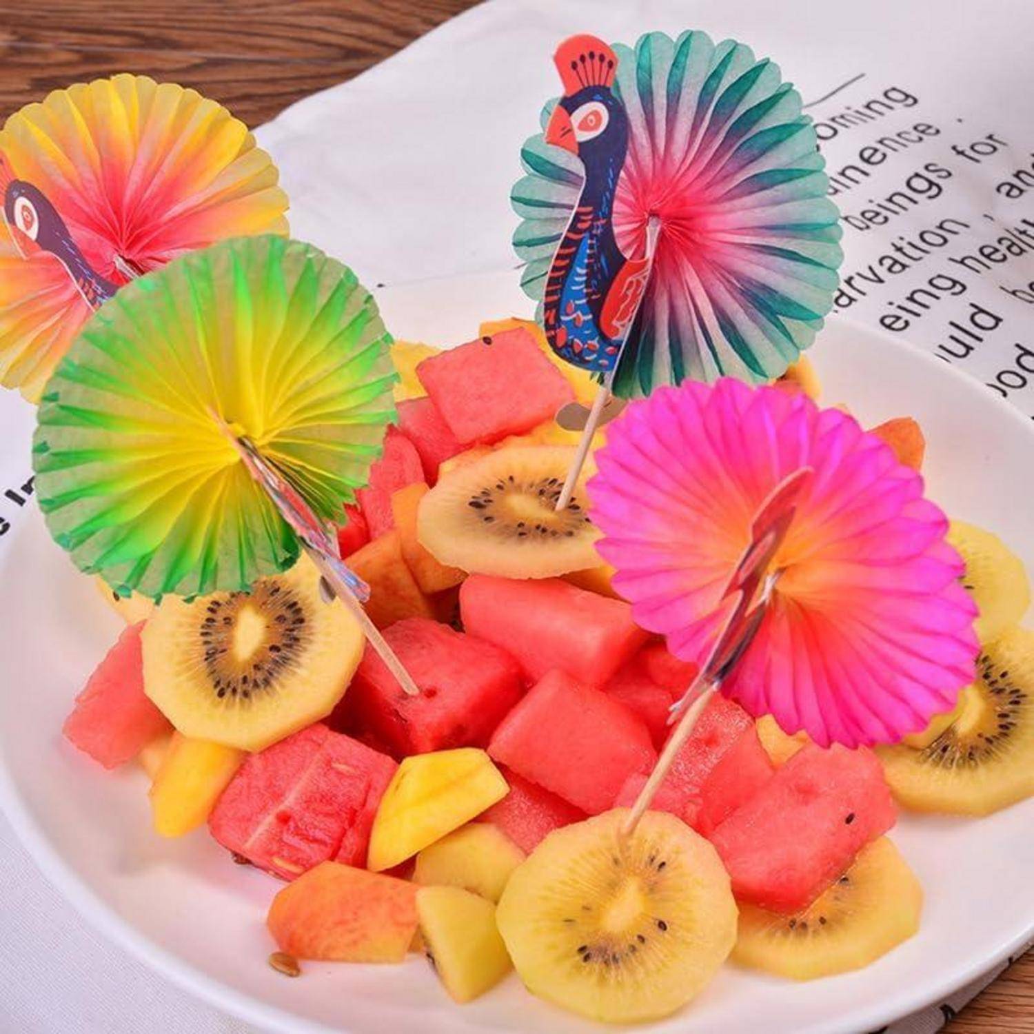 Ein bunter Obstsalat mit Wassermelonen-, Kiwi- und Mangostücken, gekrönt von dekorativen Papierfächern mit lebhaften Pfauendesigns.