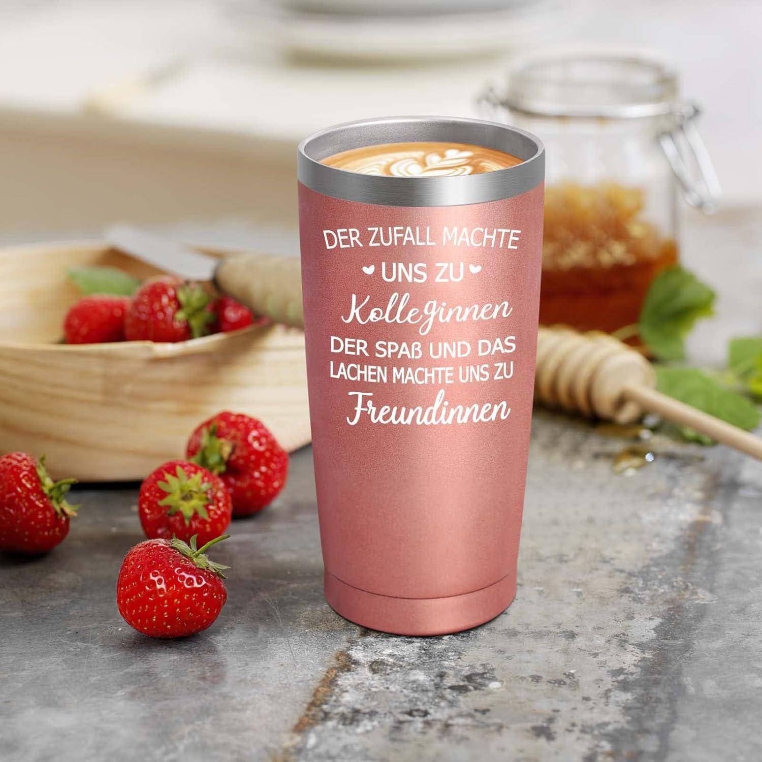 Eine rosafarbene Tasse mit deutschem Text steht auf einem Tisch neben Erdbeeren und einem Honigglas. Die Tasse enthält einen Latte mit Schaumkunst.