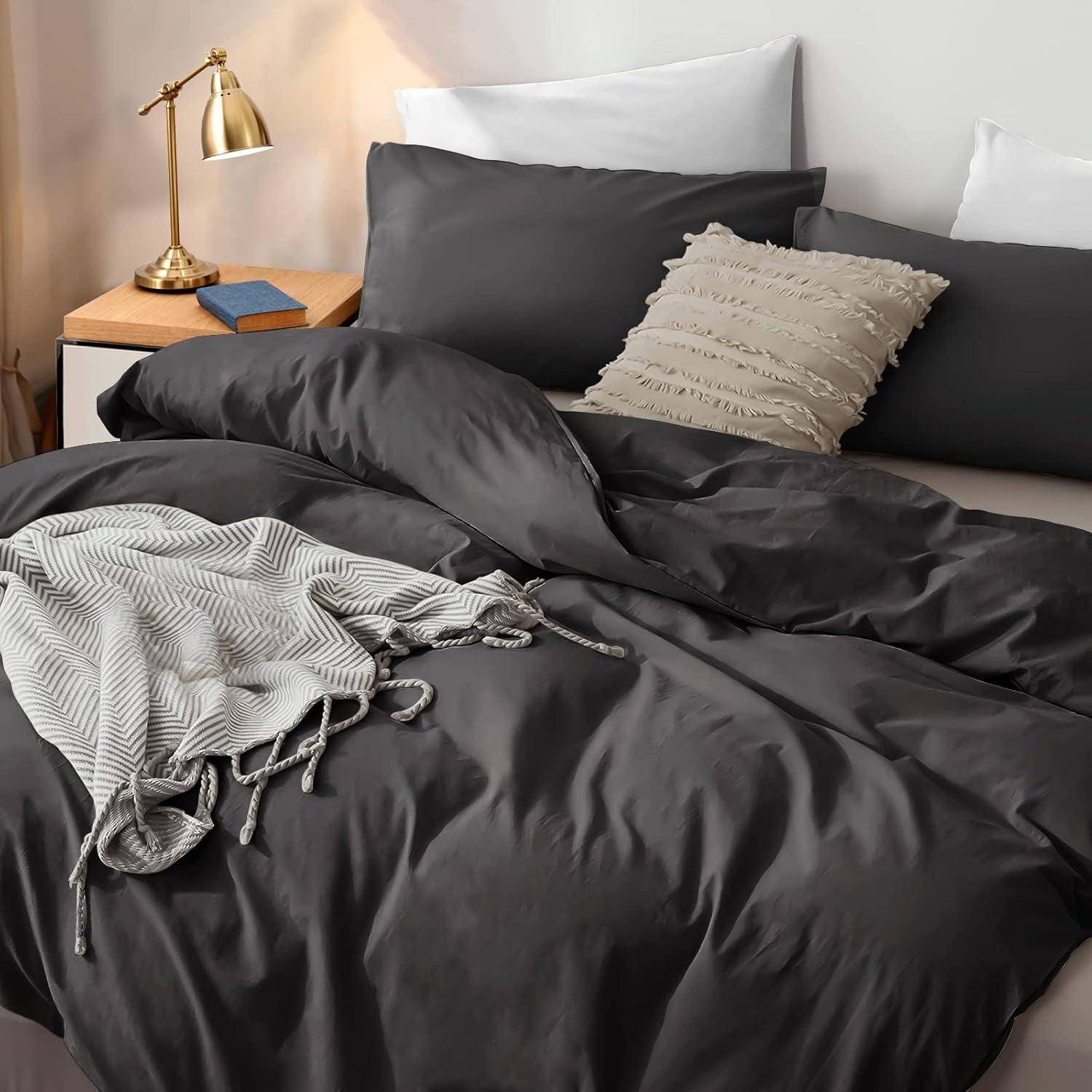Ein ordentlich gemachtes Bett mit dunkelgrauen Laken und einer gefransten Decke, neben einem Holz-Nachttisch mit einer Messinglampe und einem Buch.