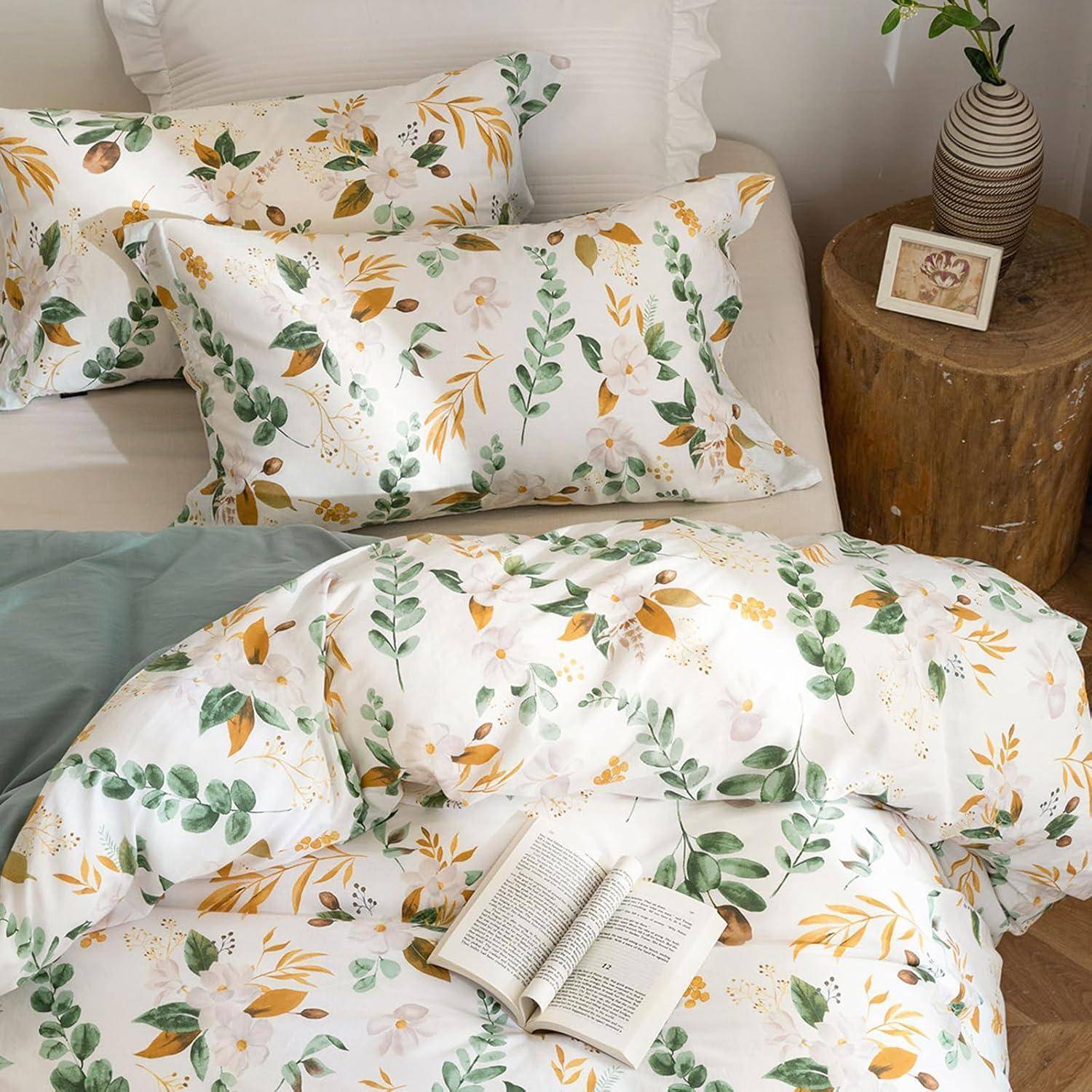 Bettwäsche-Set mit Blumenmuster umfasst Kissen und eine Bettdecke. Ein Buch liegt aufgeschlagen auf dem Bett. Ein kleiner Holztisch trägt eine Pflanze.