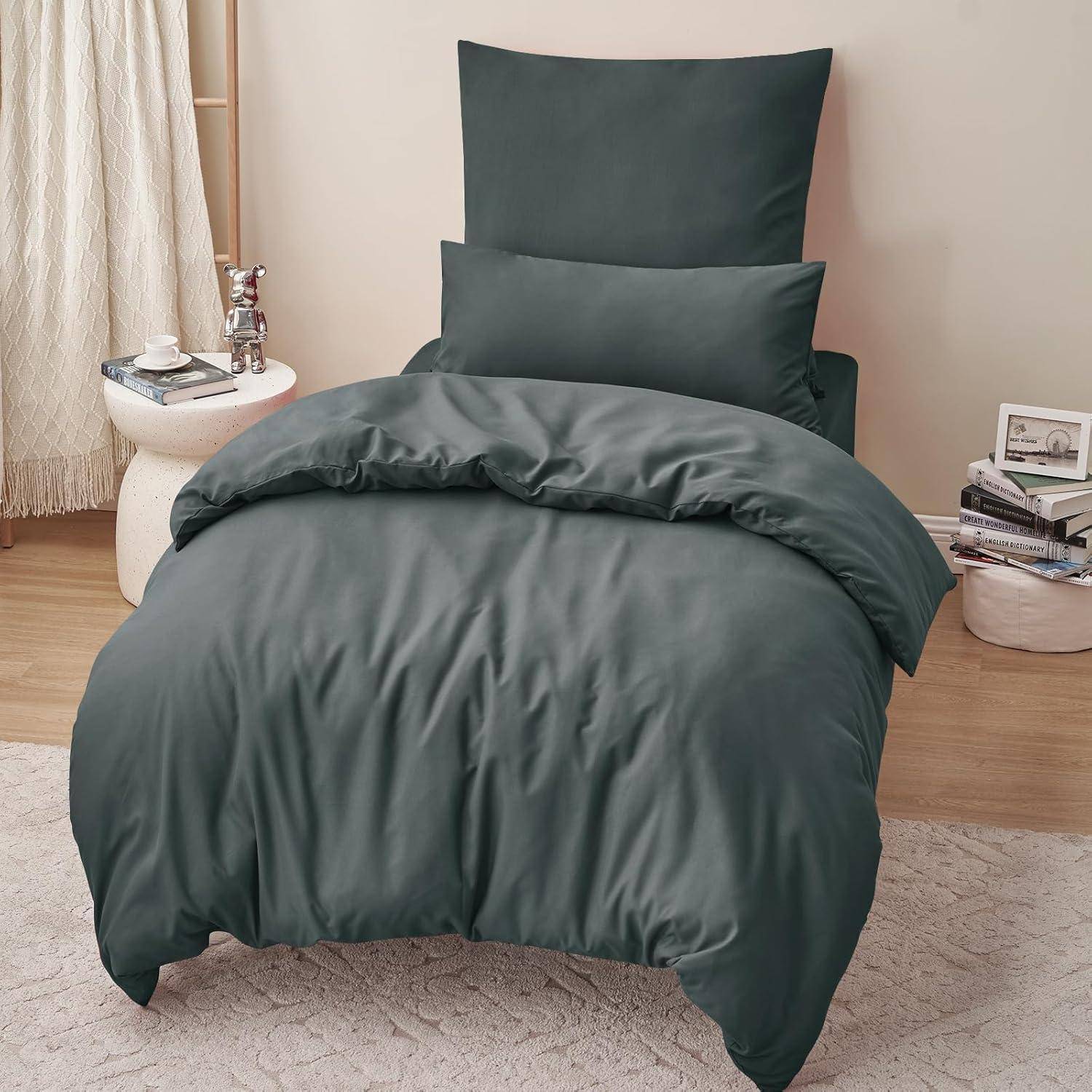 Ein ordentlich gemachtes Bett mit dunkelgrauer Bettwäsche, einschließlich Kissen, Decke und Spannbetttuch, steht neben einem kleinen Holznachttisch mit Büchern und einer Lampe.