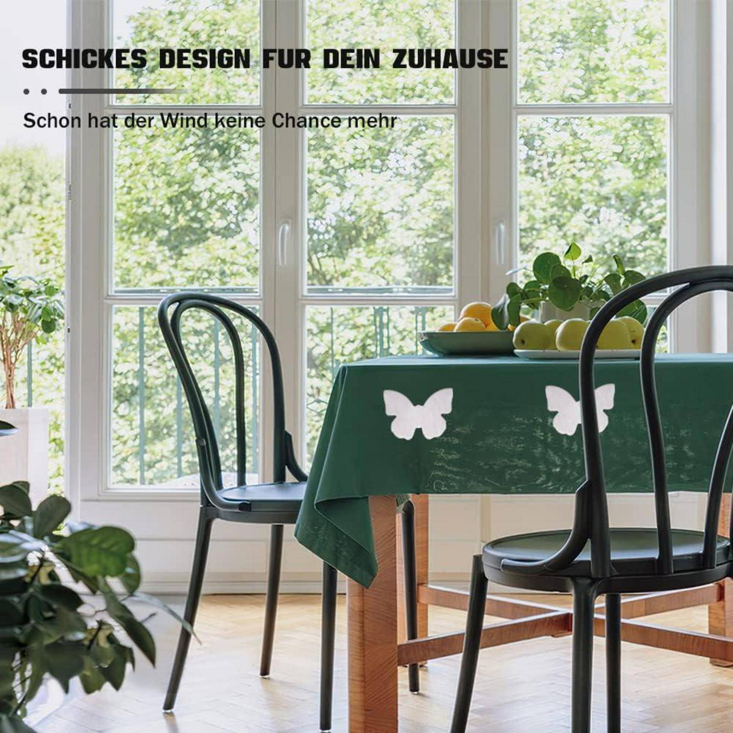 Ein Esstisch mit einer grünen Tischdecke, die weiße Schmetterlingsdesigns aufweist, ist neben zwei Stühlen in der Nähe eines großen Fensters mit Gartenblick aufgestellt. Der deutsche Text im Bild deutet auf ein stilvolles Design für das Zuhause hin.