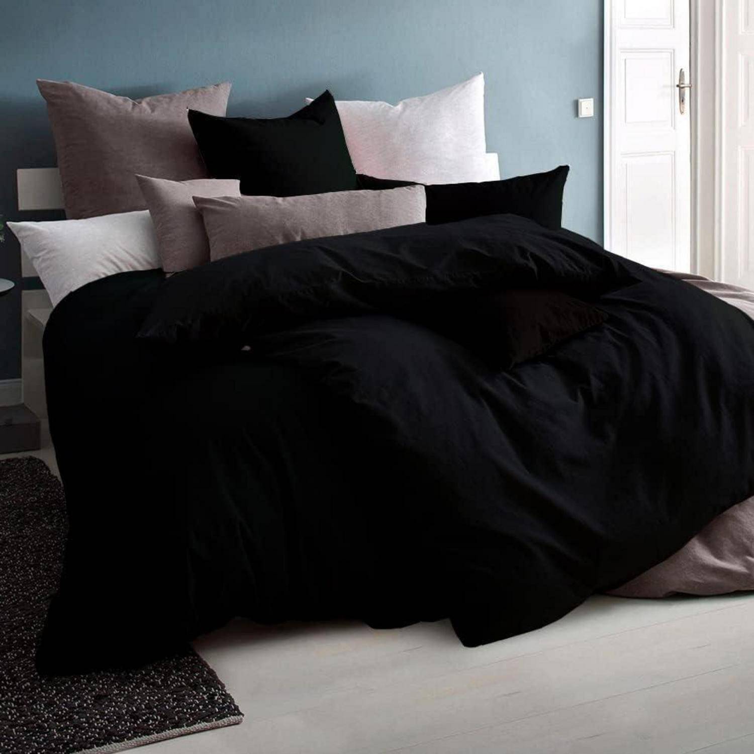 Ein Schlafzimmer mit einem ordentlich gemachten Bett, das mit schwarzen und grauen Kissen und Bettdecken bedeckt ist, vor einer blauen Wand. Eine Tür ist teilweise zu sehen.