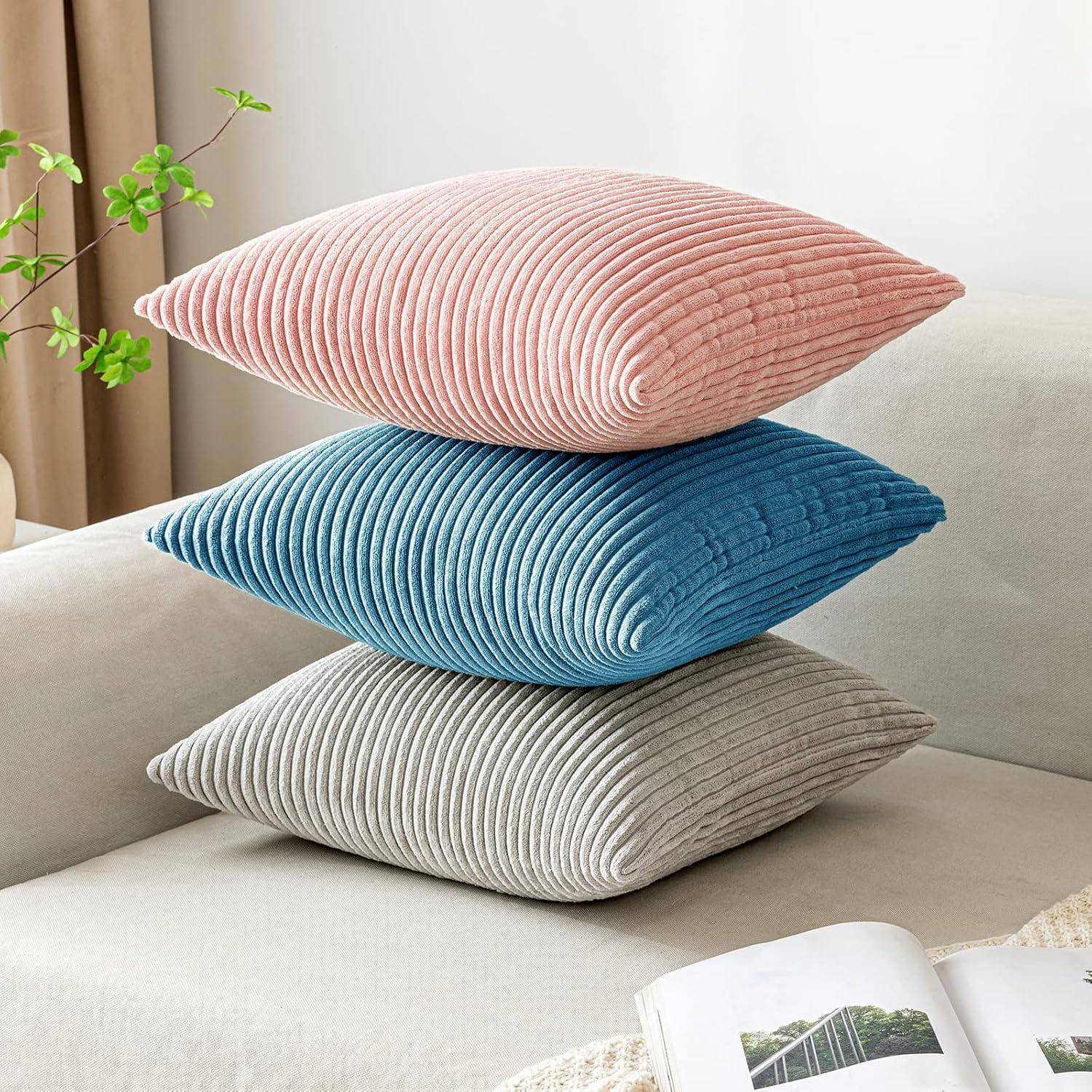 Ein Stapel von drei Cordkissen in Rosa, Blau und Grau auf einem beigen Sofa, mit einer Pflanze im Hintergrund, die eine gemütliche Atmosphäre schafft.
