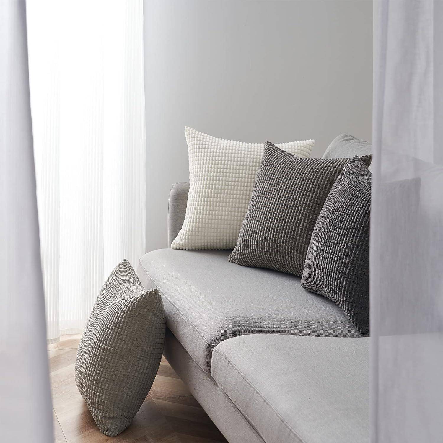 Ein hellgrauer Sofa mit verschiedenen Kissen in Strukturtexturen in Grau- und Beigetönen, platziert neben weißen transparenten Vorhängen in einem minimalistischen Raum.