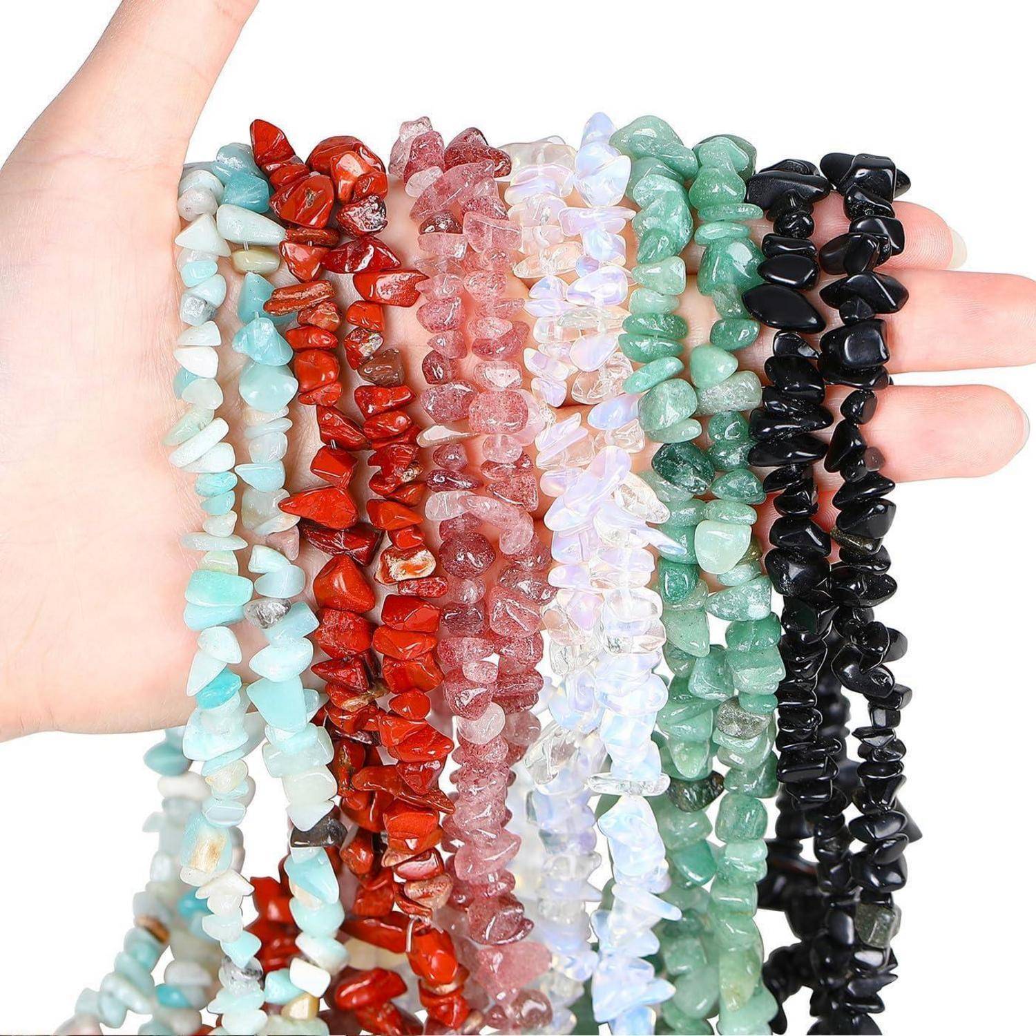 Eine Hand hält mehrere Stränge bunter Steinperlenarmbänder, darunter Türkis, Rot, Transparent, Grün und Schwarz, vor einem weißen Hintergrund.