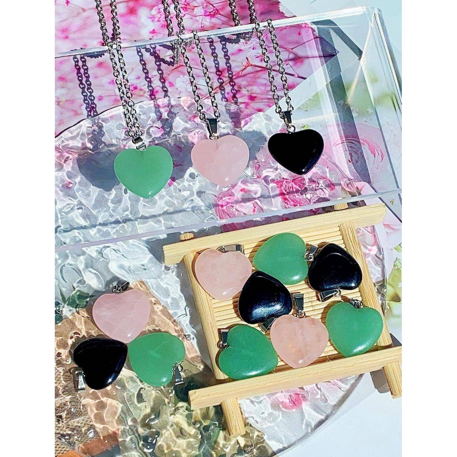 Drei herzförmige Halsketten und sechs passende Anhänger in Grün, Rosa und Schwarz, die auf einem Bambusgestell vor einem Blumenhintergrund ausgestellt sind.