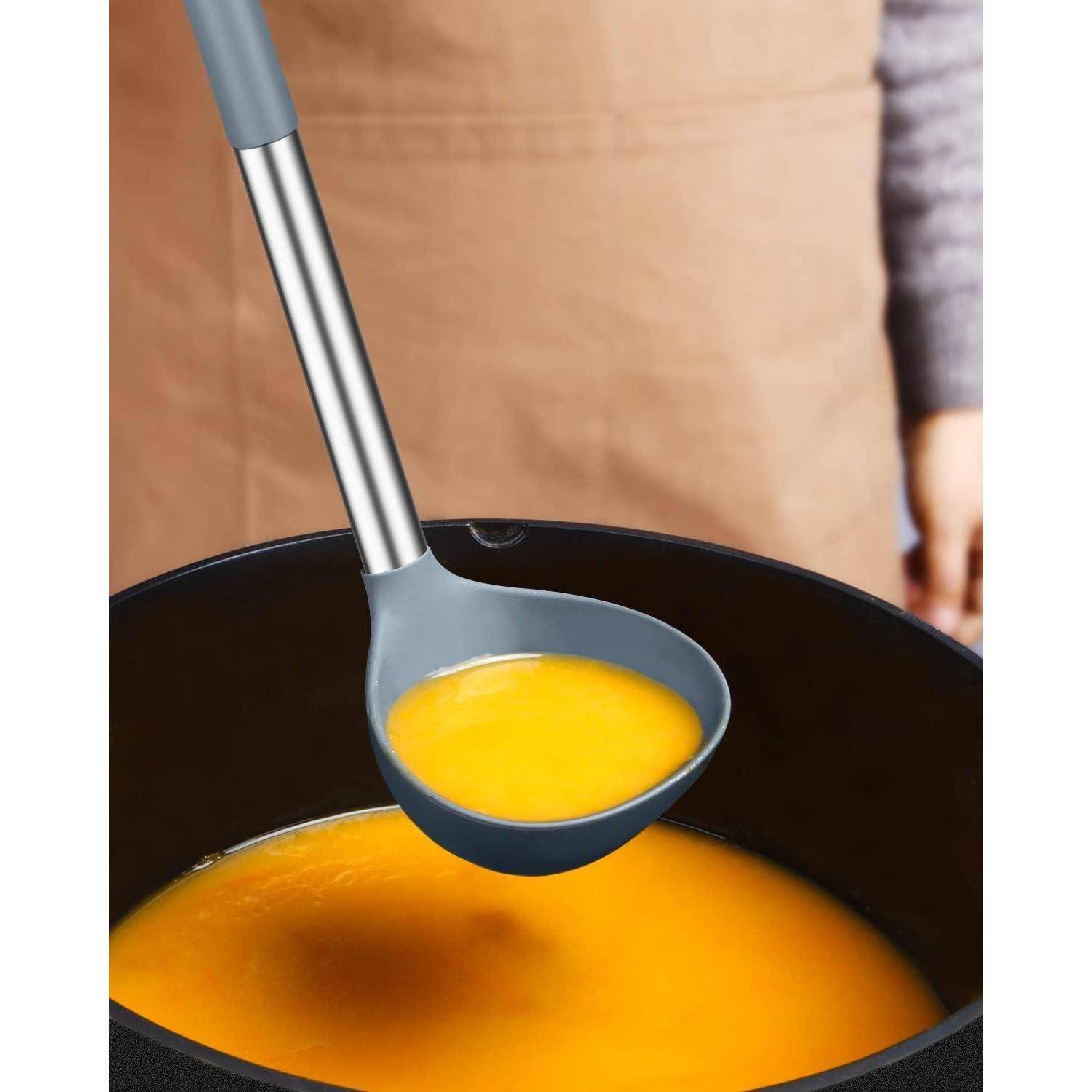 Eine Person hält einen Löffel aus Metall und Silikon über eine Schüssel mit leuchtend orangefarbener Suppe, bereit zum Servieren. Der Hintergrund zeigt einen unscharfen Schurz.