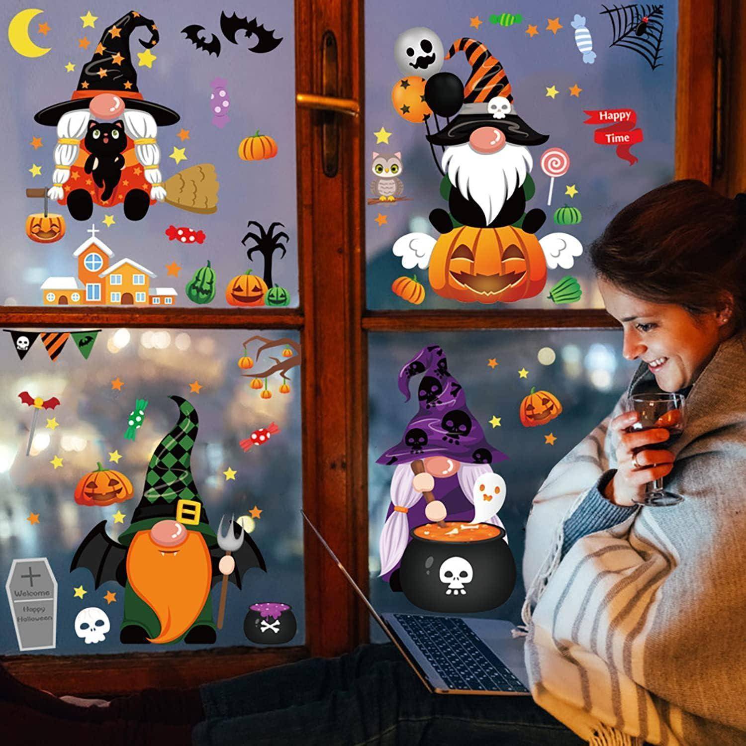 Eine Frau, in eine Decke gehüllt, sitzt neben einem Fenster, das mit Halloween-Stickern geschmückt ist, darunter Gnome, Kürbisse und Fledermäuse.