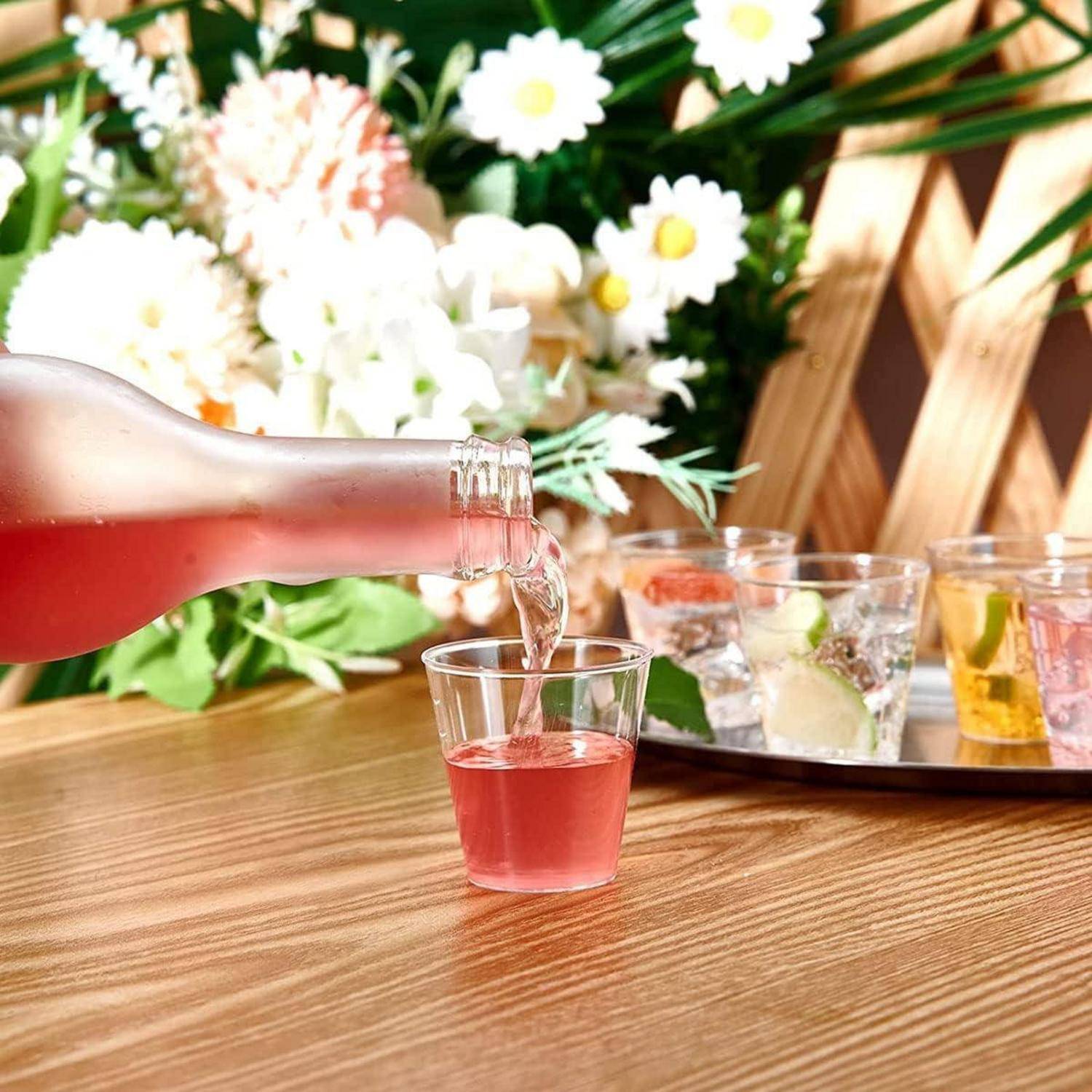 Eine Nahaufnahme eines rosa Getränks, das in ein kleines Glas auf einem Holztisch gegossen wird, mit einem Blumenarrangement und einem Tablett mit Getränken im Hintergrund.