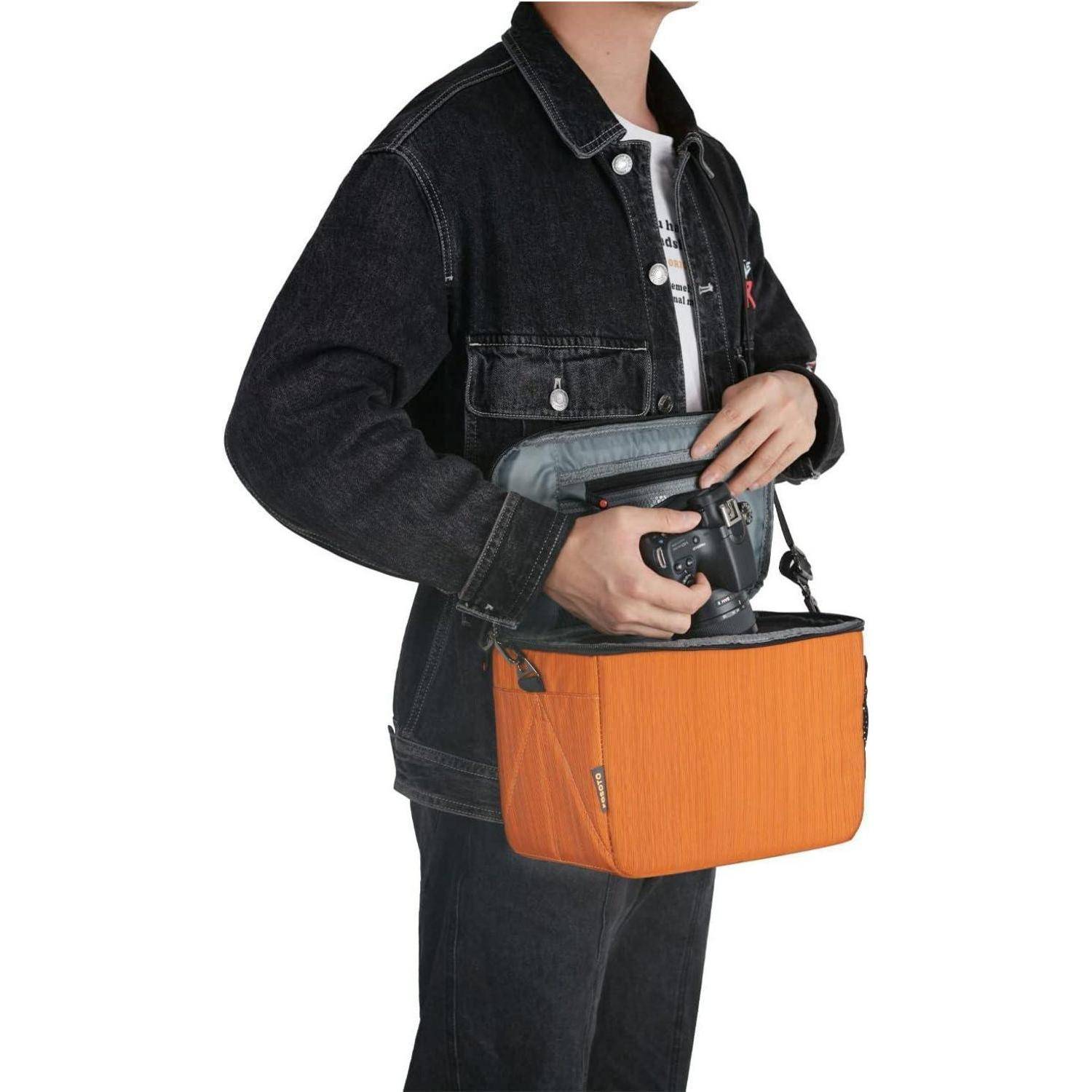 Eine Person hält eine orangefarbene Tasche mit einem schwarzen Deckel und trägt eine schwarze Jeansjacke. Die Szene deutet auf Reise oder Organisation hin.