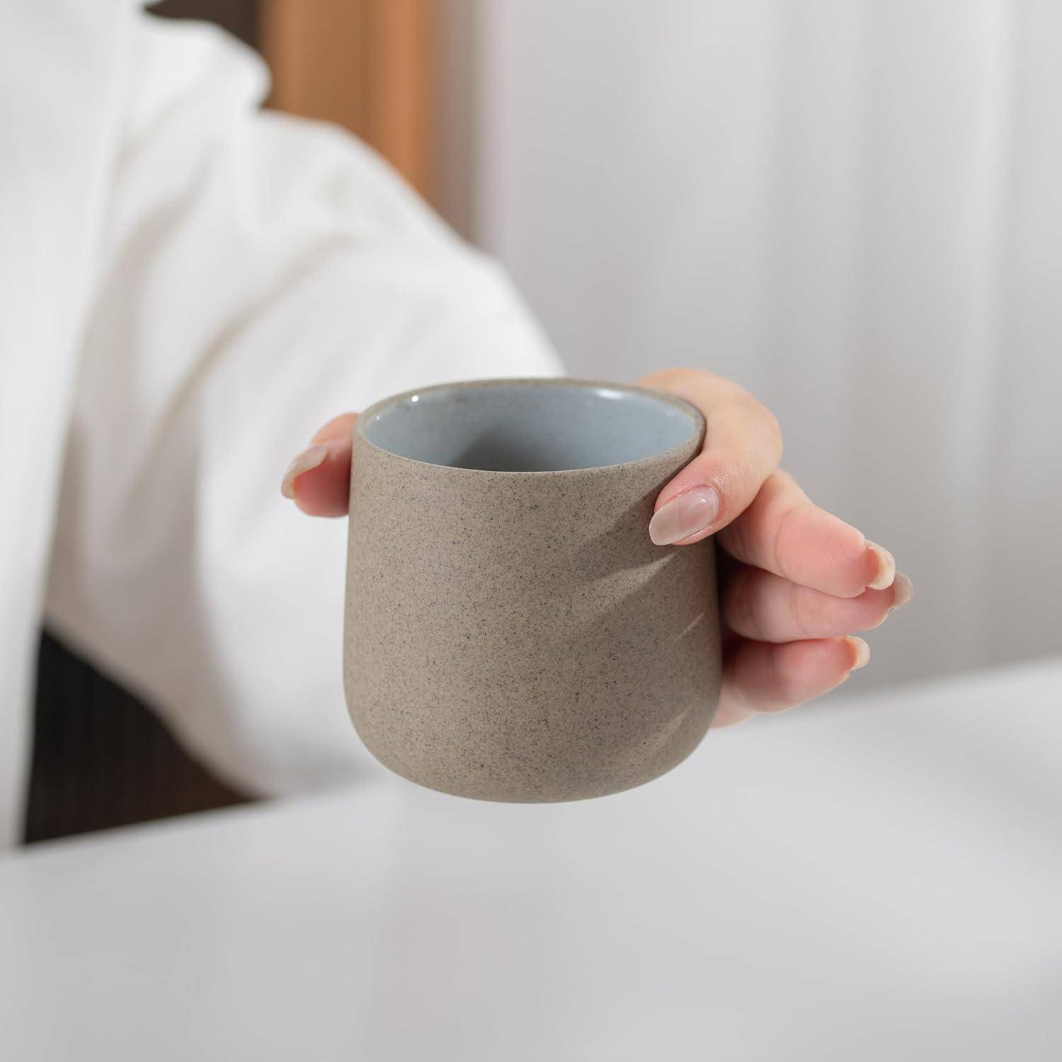 Eine Nahaufnahme einer Person, die eine kleine, beige Keramiktasse mit beiden Händen hält, über einer weißen Tischoberfläche positioniert.
