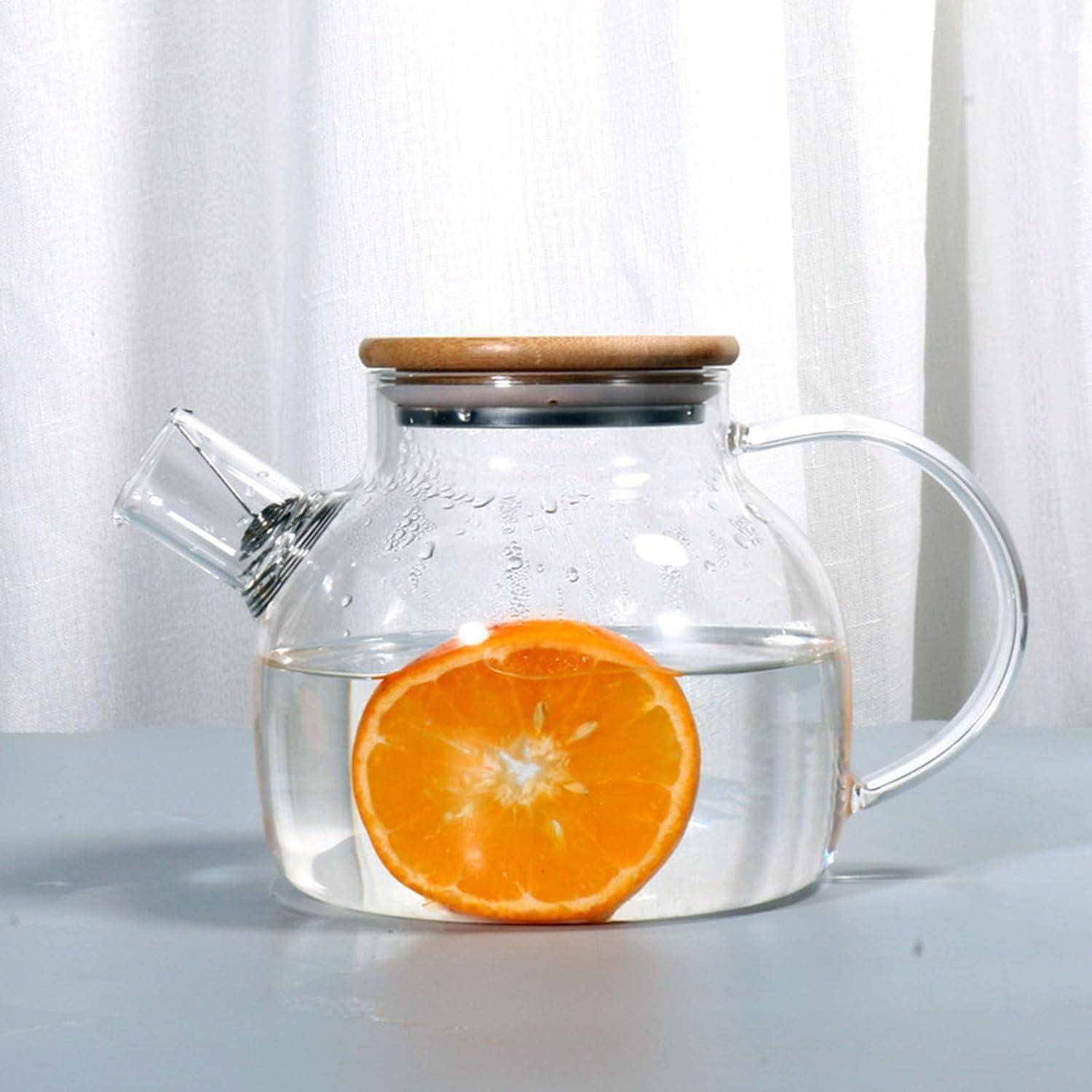 Eine durchsichtige Teekanne, gefüllt mit Wasser, enthält eine Scheibe Orange, die auf einer hellblauen Oberfläche vor einem weißen Vorhanghintergrund steht.