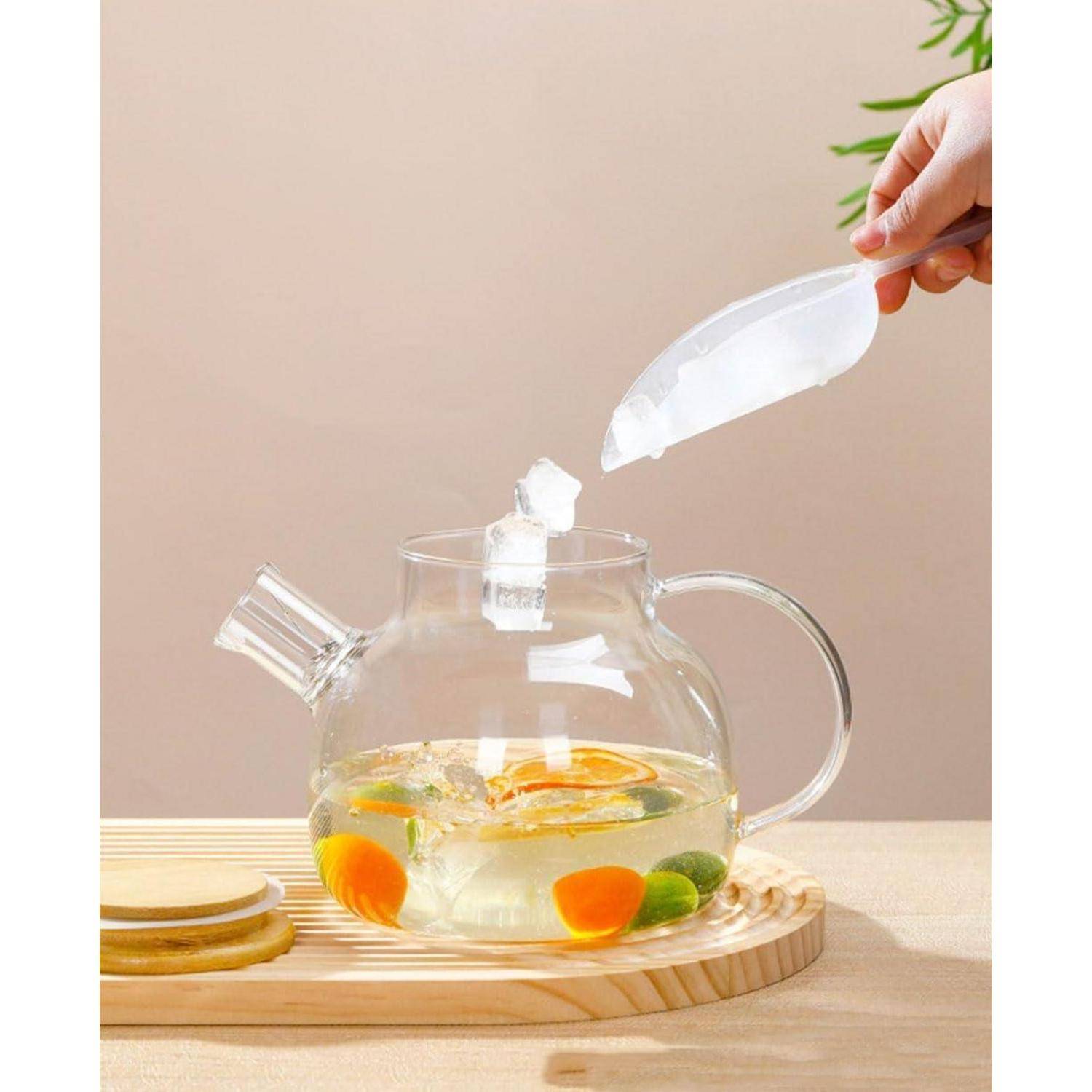 Eine Glaskanne enthält Zitrusfrüchtescheiben und Kräuter, die auf einem Holztablett platziert sind. Eine Hand hält einen Eislöffel darüber und gibt Eis in die Kanne.