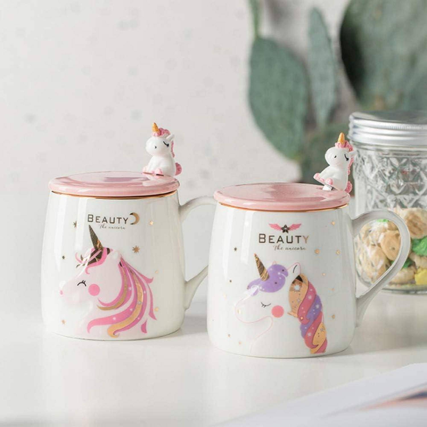 Zwei weiße Tassen mit Einhorn-Designs und rosa Deckeln, mit kleinen Einhornfiguren oben drauf. Text auf den Tassen lautet 'Beauty'.